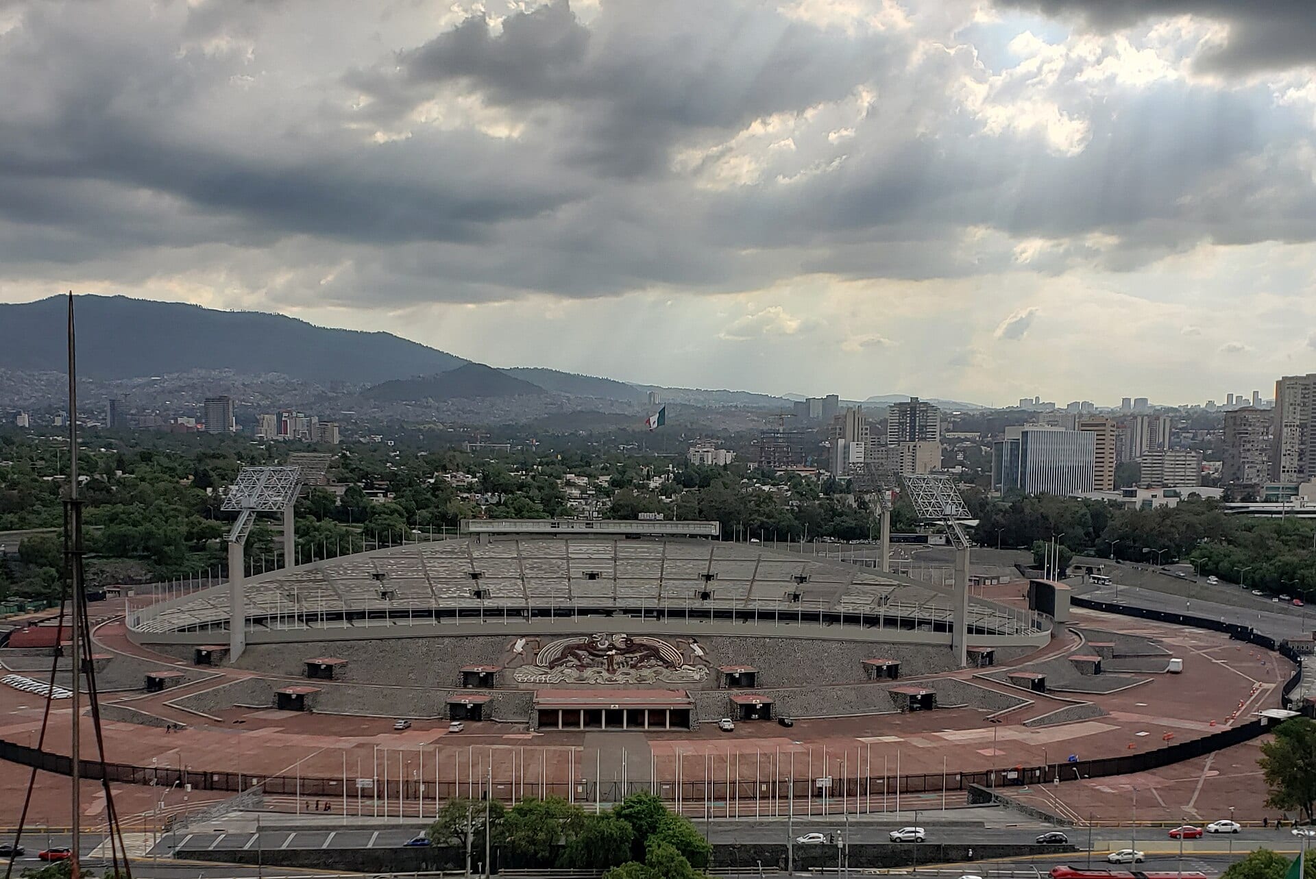 Vista elevada del Estadio Olímpico Universitario, con sus gradas vacías y el mural de la fachada, y las montañas del sur de la ciudad al fondo.
