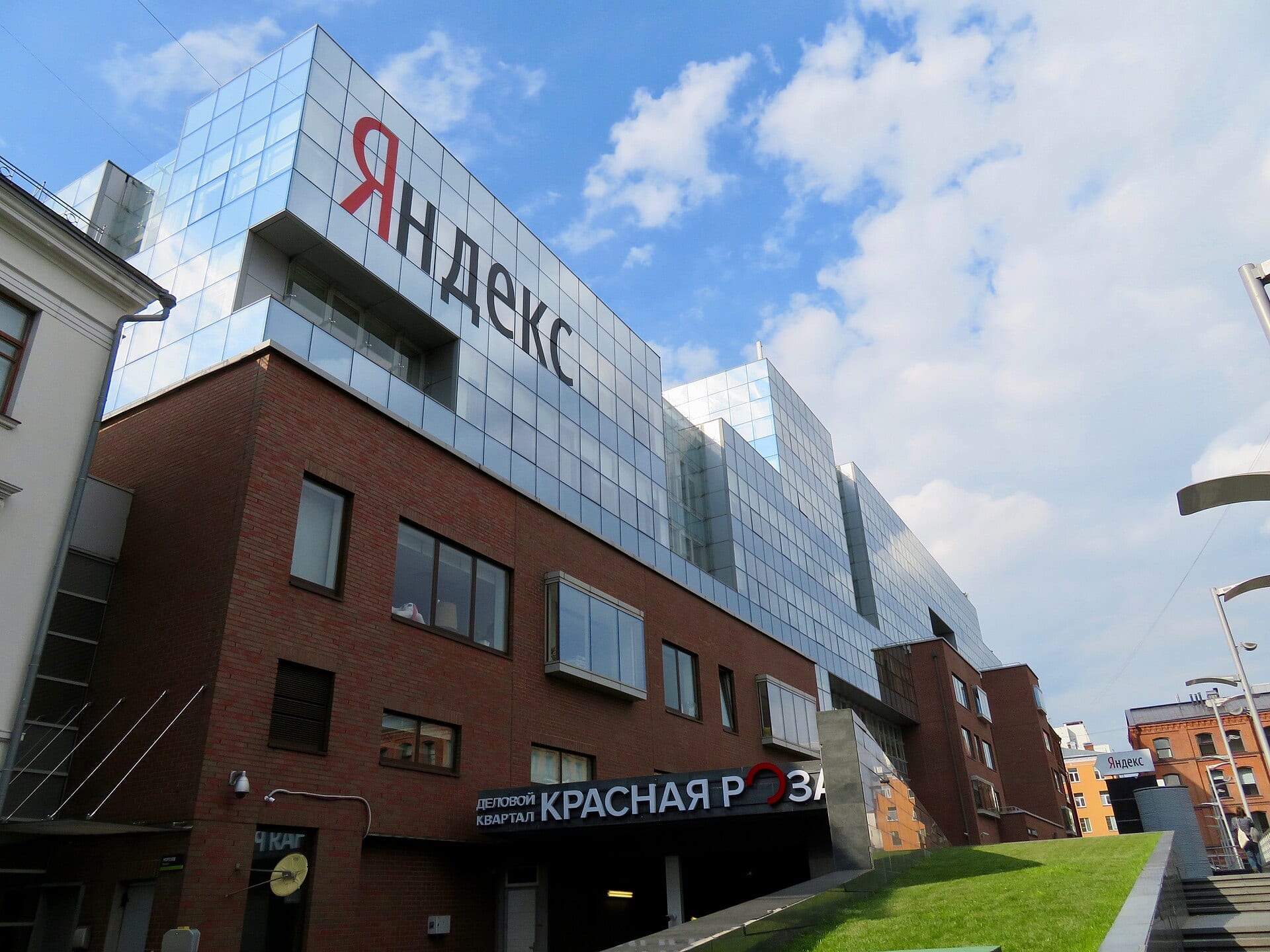 Fachada de vidrio y ladrillo de un edificio de oficinas con el logotipo rojo y negro de Yandex en letras cirílicas, bajo un cielo con nubes.