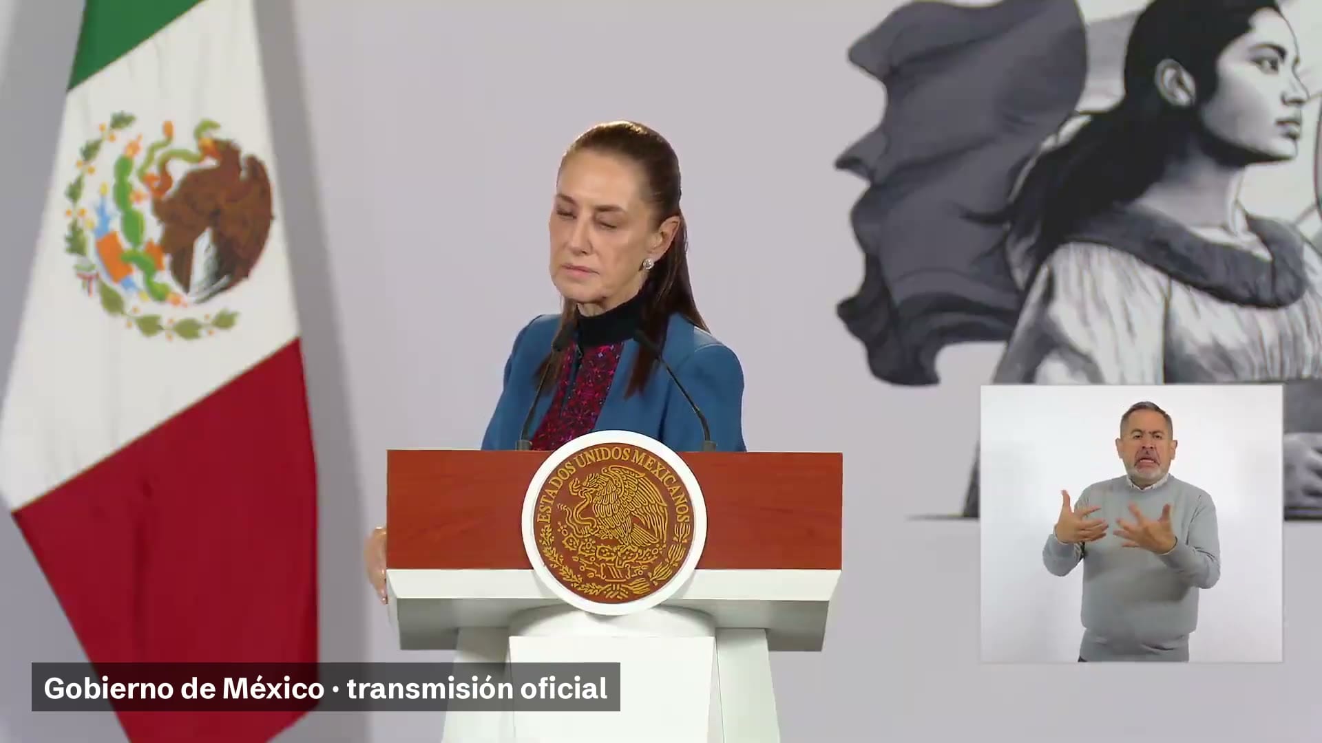 La presidenta Claudia Sheinbaum, de pie tras el podio de la conferencia matutina, con la bandera de México a su izquierda y una intérprete de lengua de señas a su derecha.
