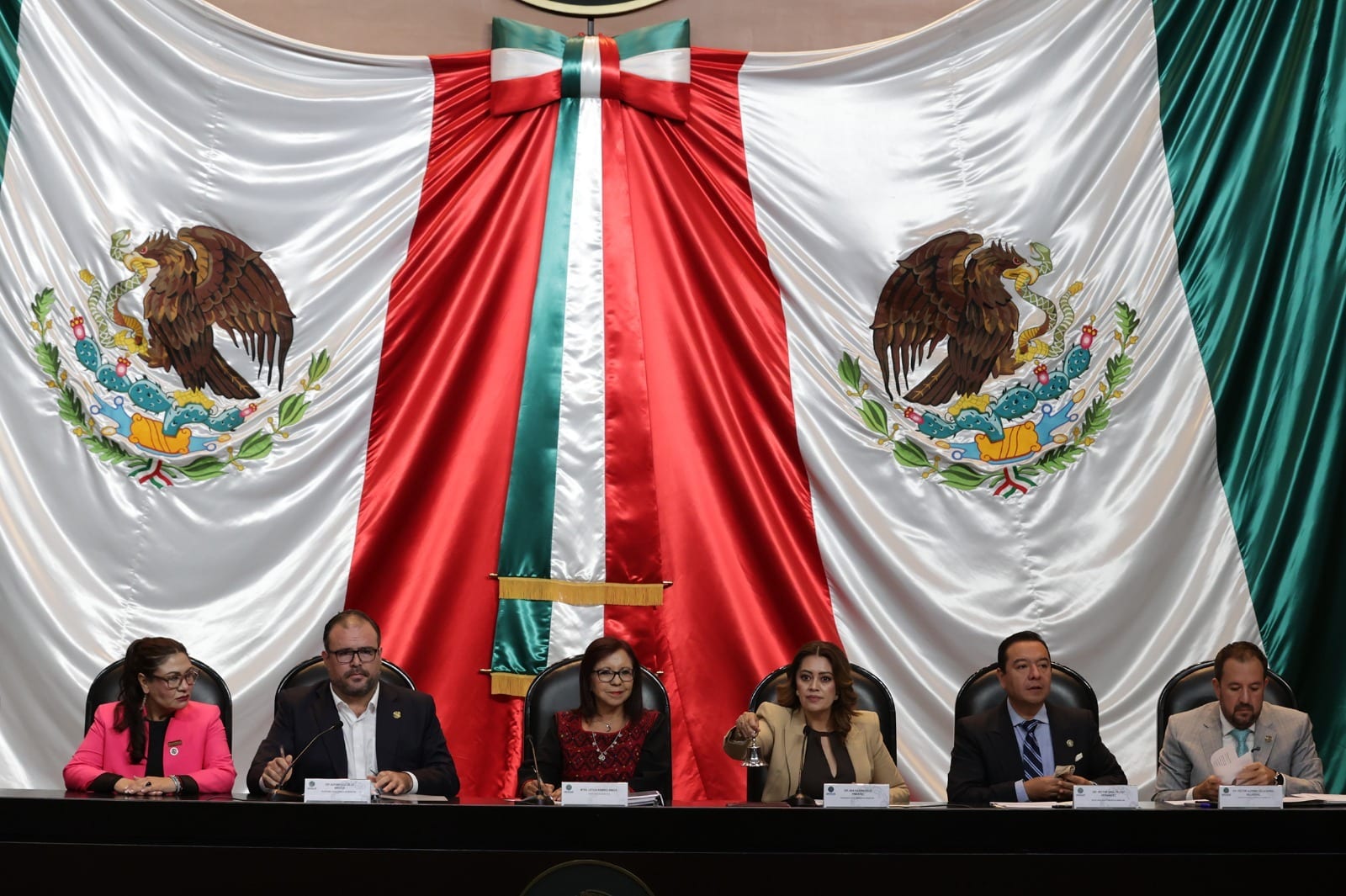 Legisladores y la secretaria de Bienestar, sentados en fila ante la mesa de sesiones; una mujer agita una campana bajo un gran lienzo con la bandera nacional.