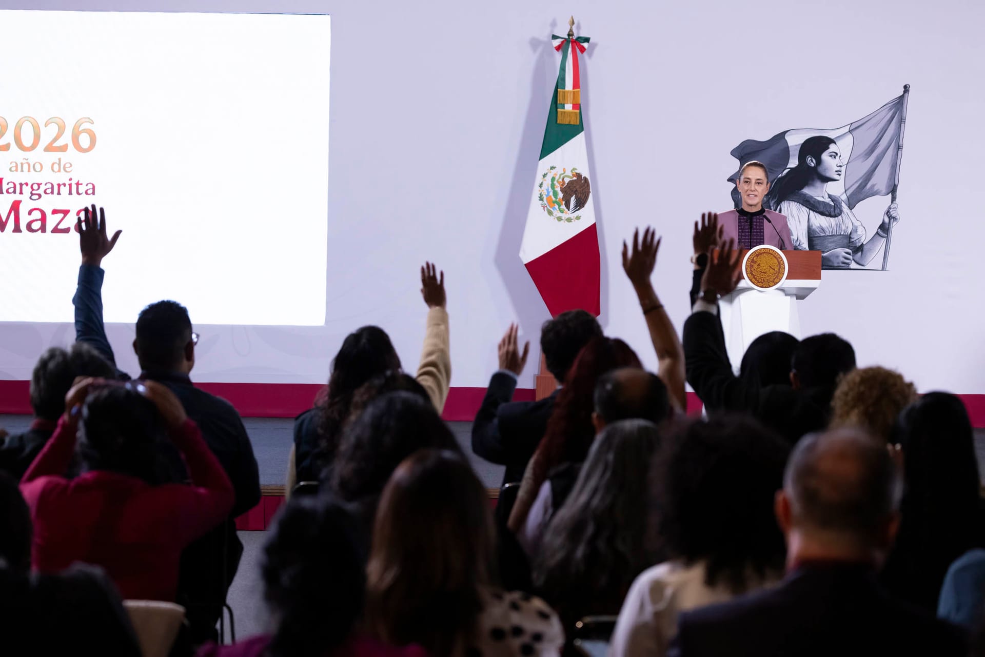 La presidenta Sheinbaum habla desde un podio con escudo nacional junto a una bandera de México, mientras reporteros levantan la mano en primer plano.