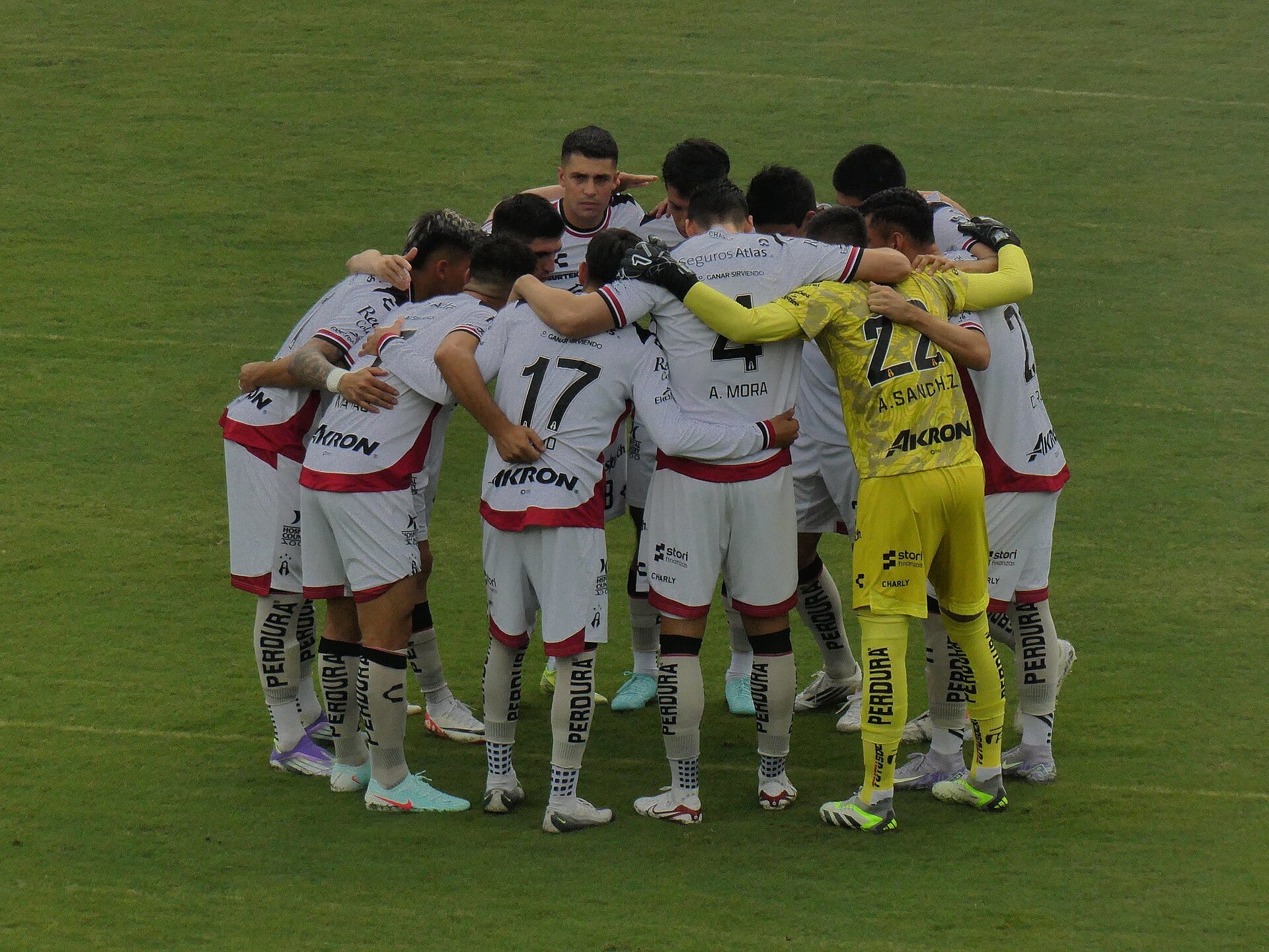 Jugadores de Atlas con camiseta blanca y detalles rojos se abrazan en círculo sobre el césped, con el portero de amarillo entre ellos.