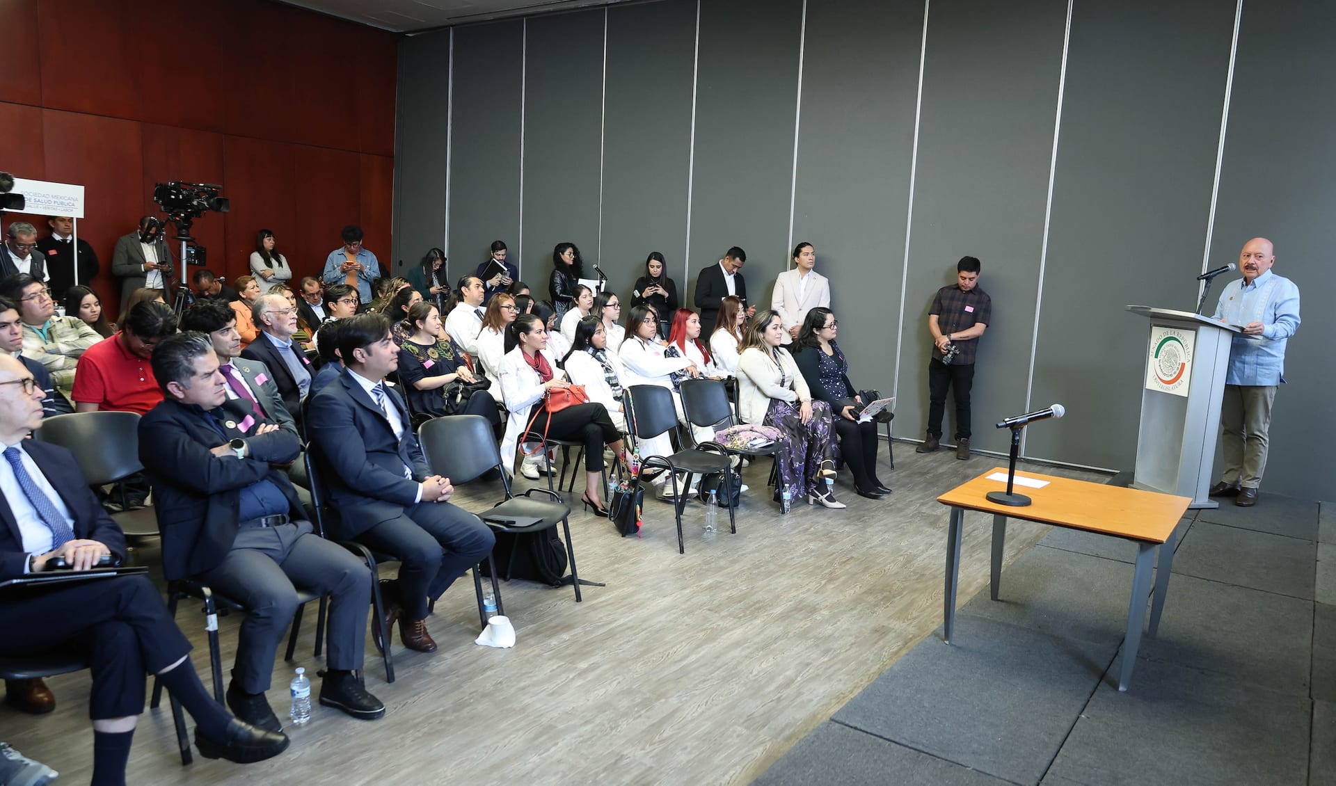 Asistentes al foro en el Senado escuchan a un orador en el atril, con el escudo nacional al frente y un cartel de la Sociedad Mexicana de Salud Pública a la izquierda.