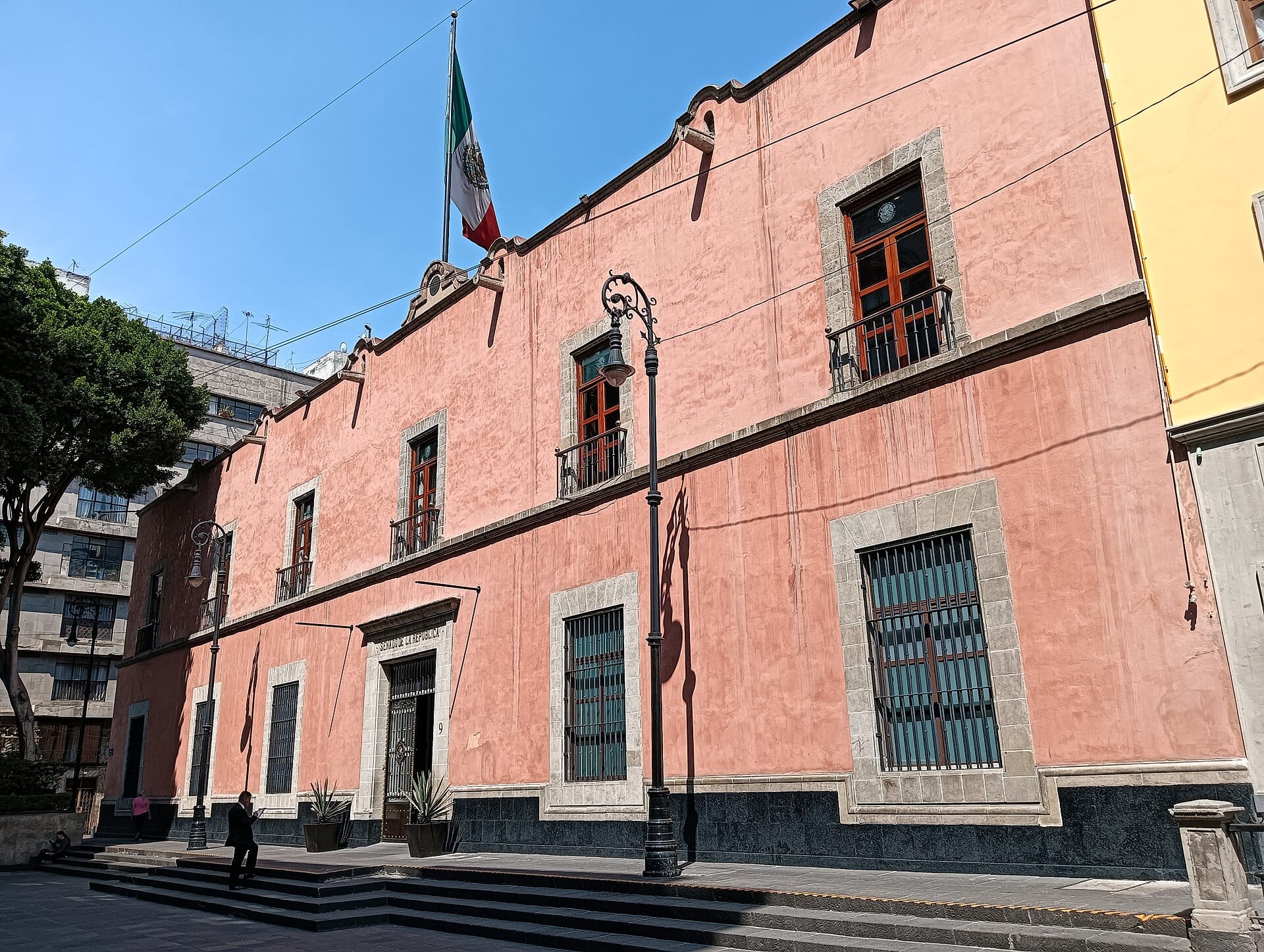 Fachada rosa de dos plantas de una casona histórica con una bandera de México en lo alto y la placa Senado de la República sobre la puerta.