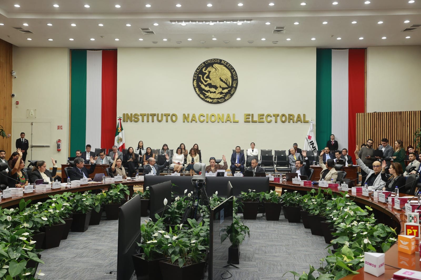 Integrantes del Consejo General del INE levantan la mano en la sala de sesiones, frente al escudo nacional y el letrero del Instituto Nacional Electoral.