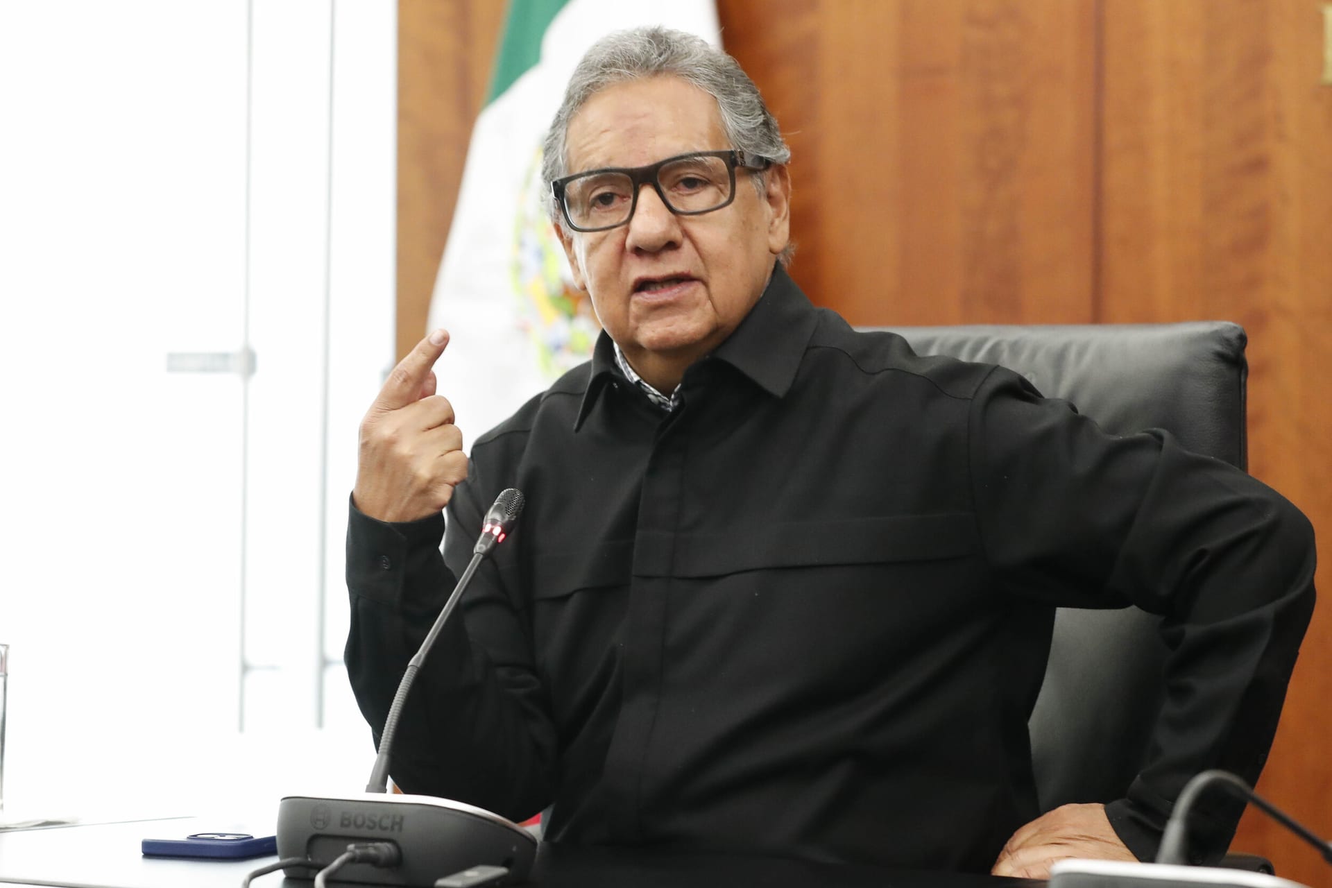 Higinio Martínez Miranda, con lentes y camisa negra, habla frente a un micrófono junto a una bandera de México y levanta el índice de la mano izquierda.