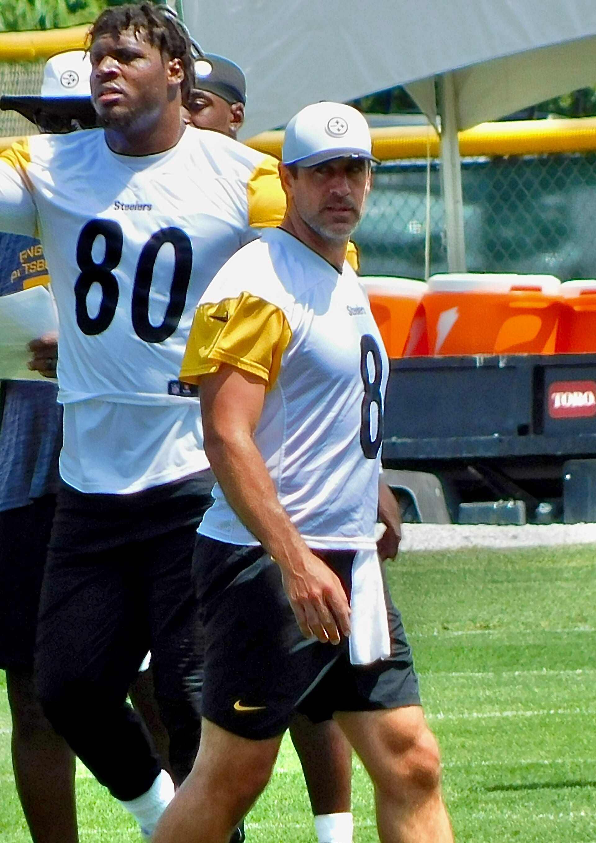 Aaron Rodgers, wearing a cap and a Steelers number 8 practice jersey, walks across a training field.