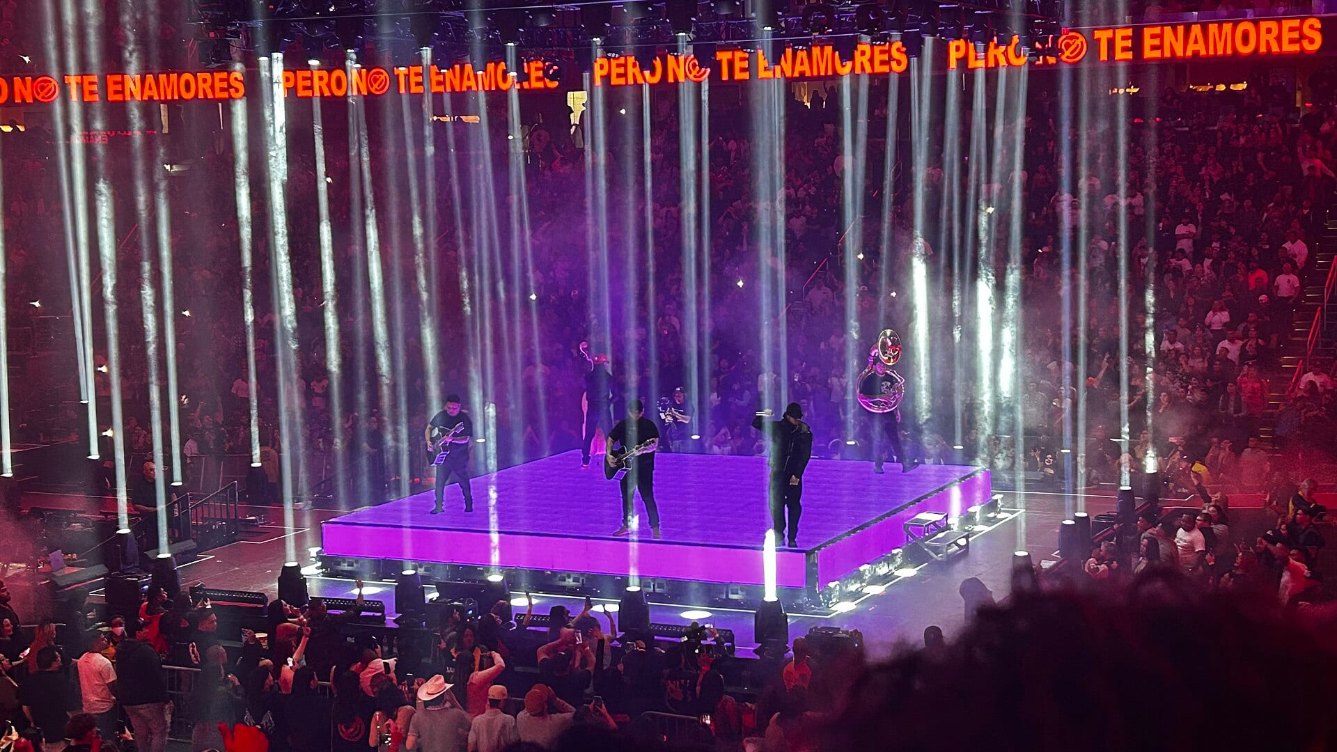 Cuatro integrantes de Fuerza Regida tocan sobre una tarima morada rodeada de haces de luz, con el público alrededor, en un concierto de 2024 en Washington.