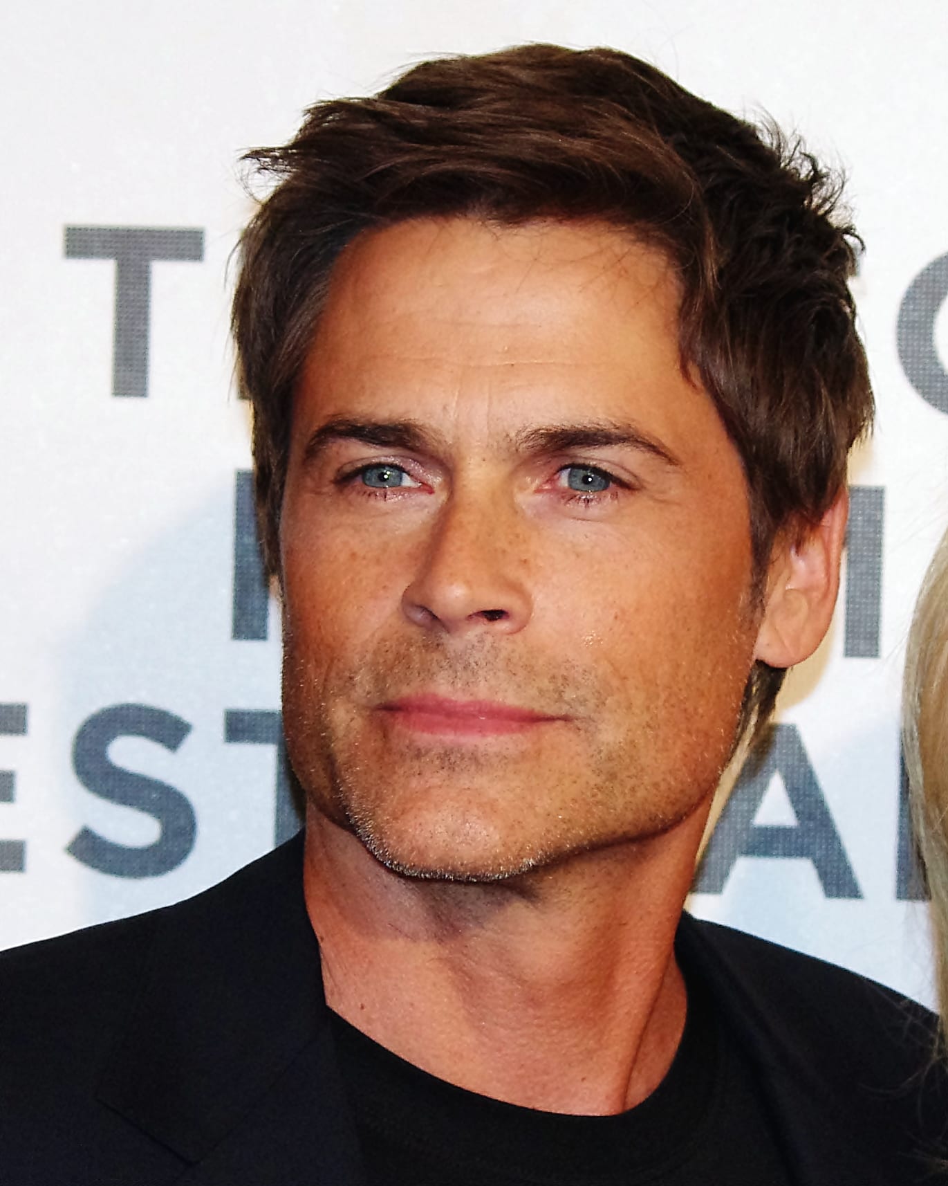Retrato de Rob Lowe, de cabello castaño y barba corta, con saco oscuro y playera negra frente a un fondo blanco con letras grises.