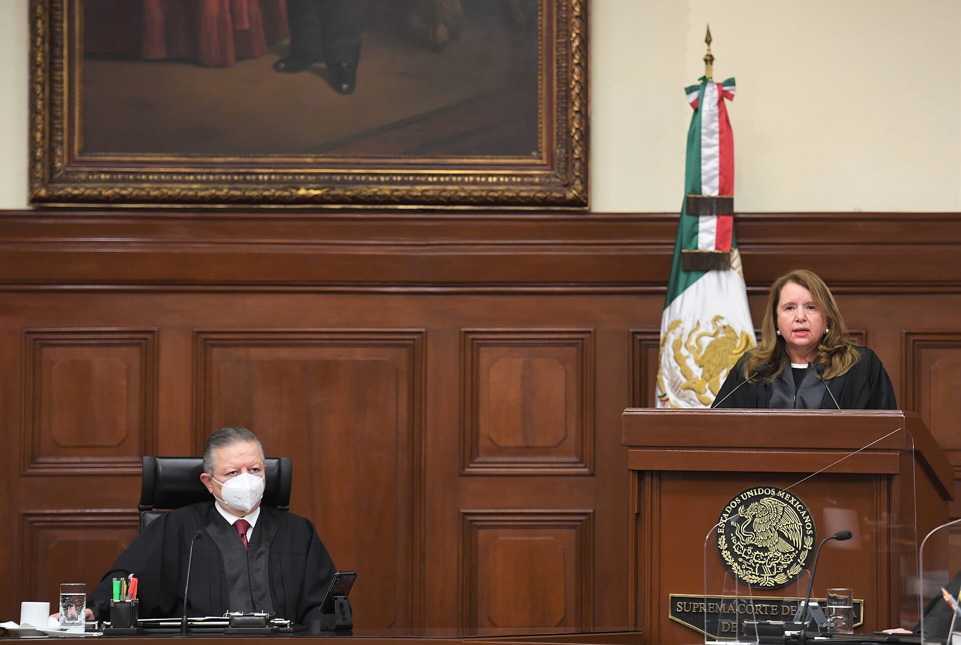 Una ministra habla desde el estrado del Pleno de la Corte, junto a una bandera de México, mientras un ministro con cubrebocas la escucha sentado.