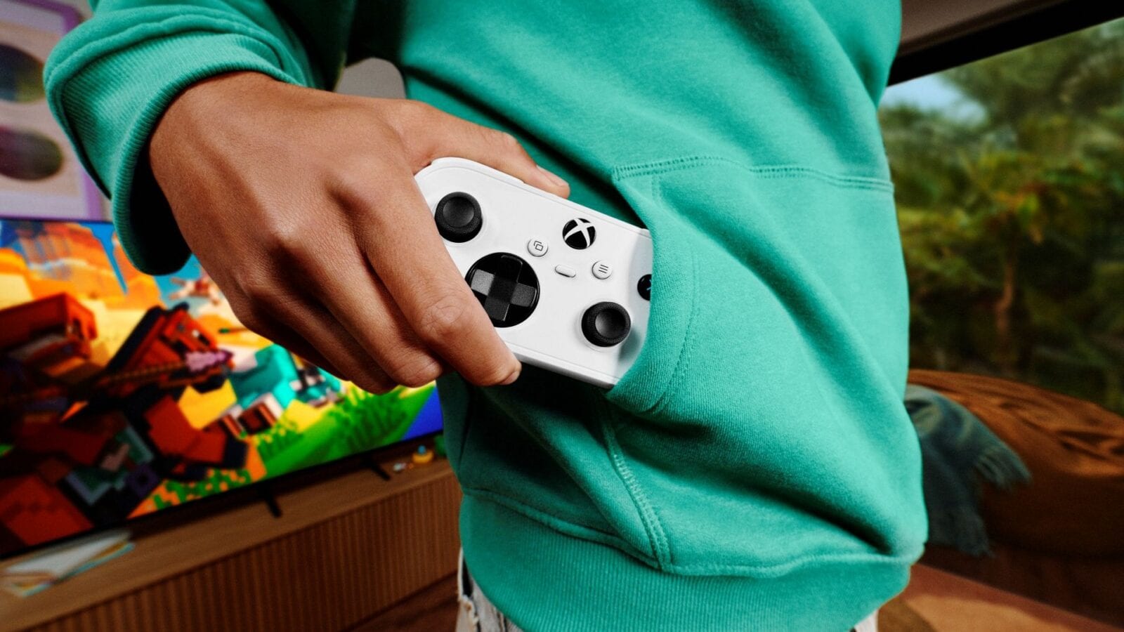 A person in a green hoodie slips a compact white Xbox controller into the pocket, with a game on a screen behind.