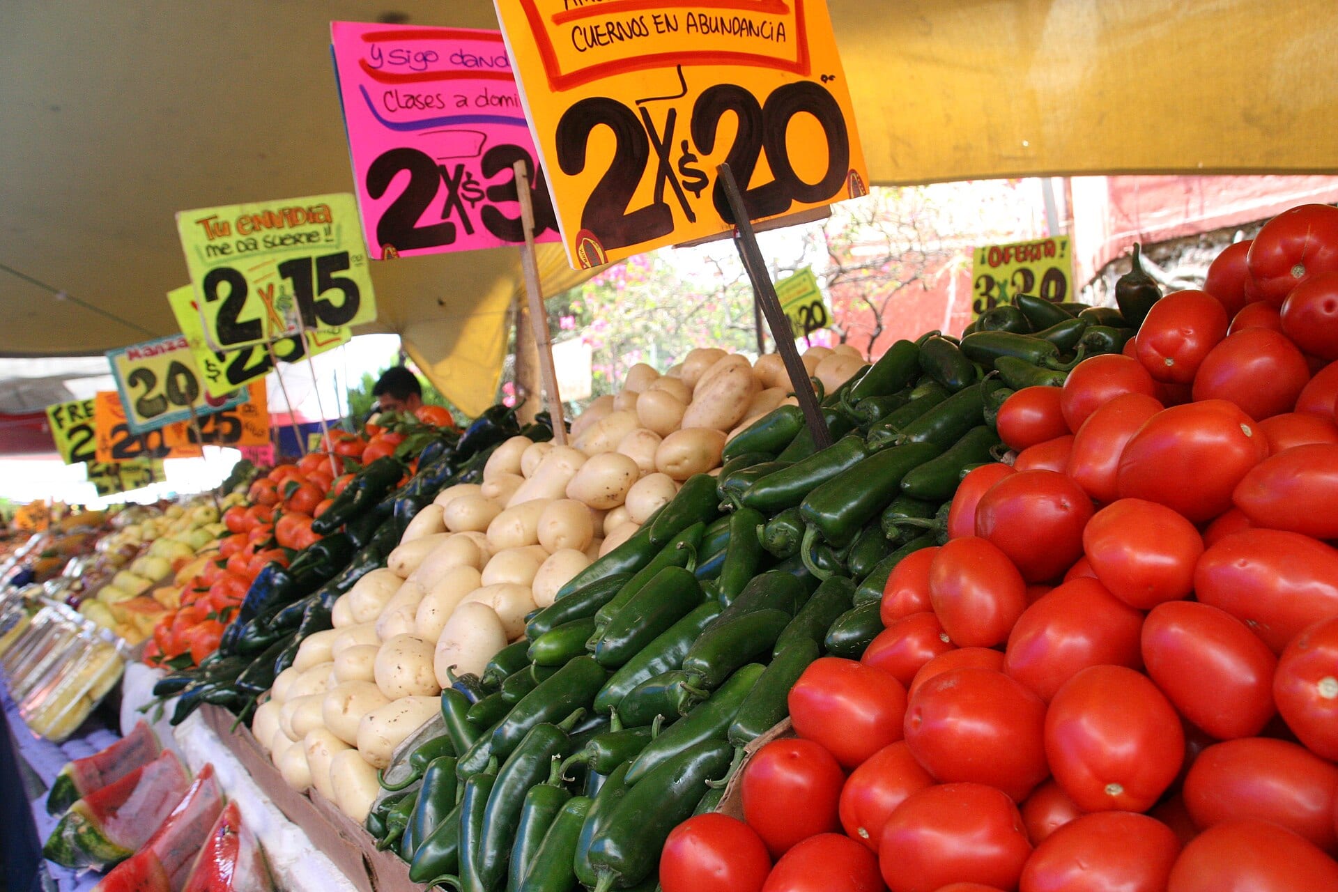 Puesto de mercado con jitomates, chiles verdes y papas apilados bajo letreros de colores que anuncian dos piezas por veinte pesos.
