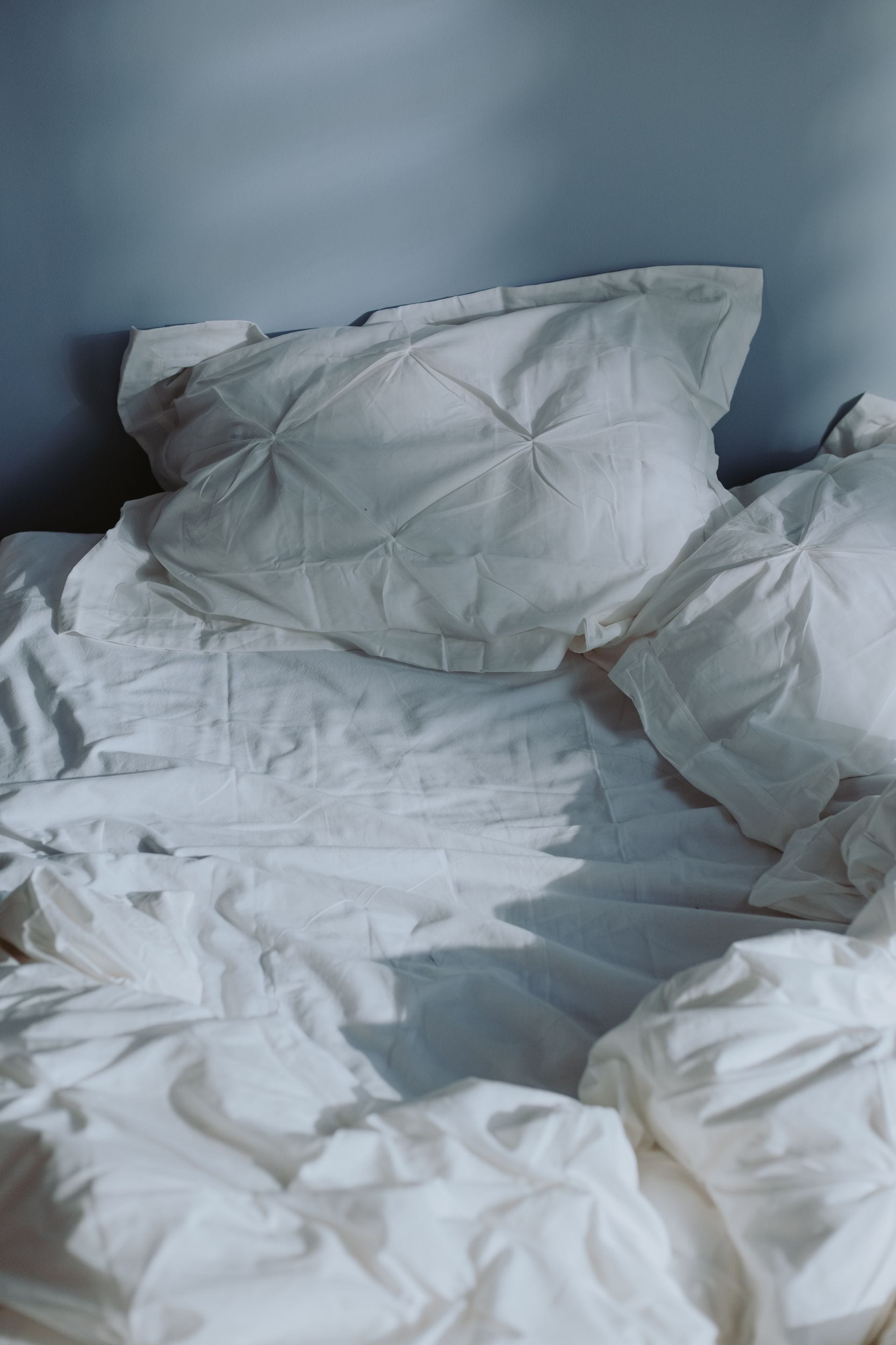 Cama deshecha con sábanas y almohadas blancas arrugadas frente a una pared azul, bajo luz suave.