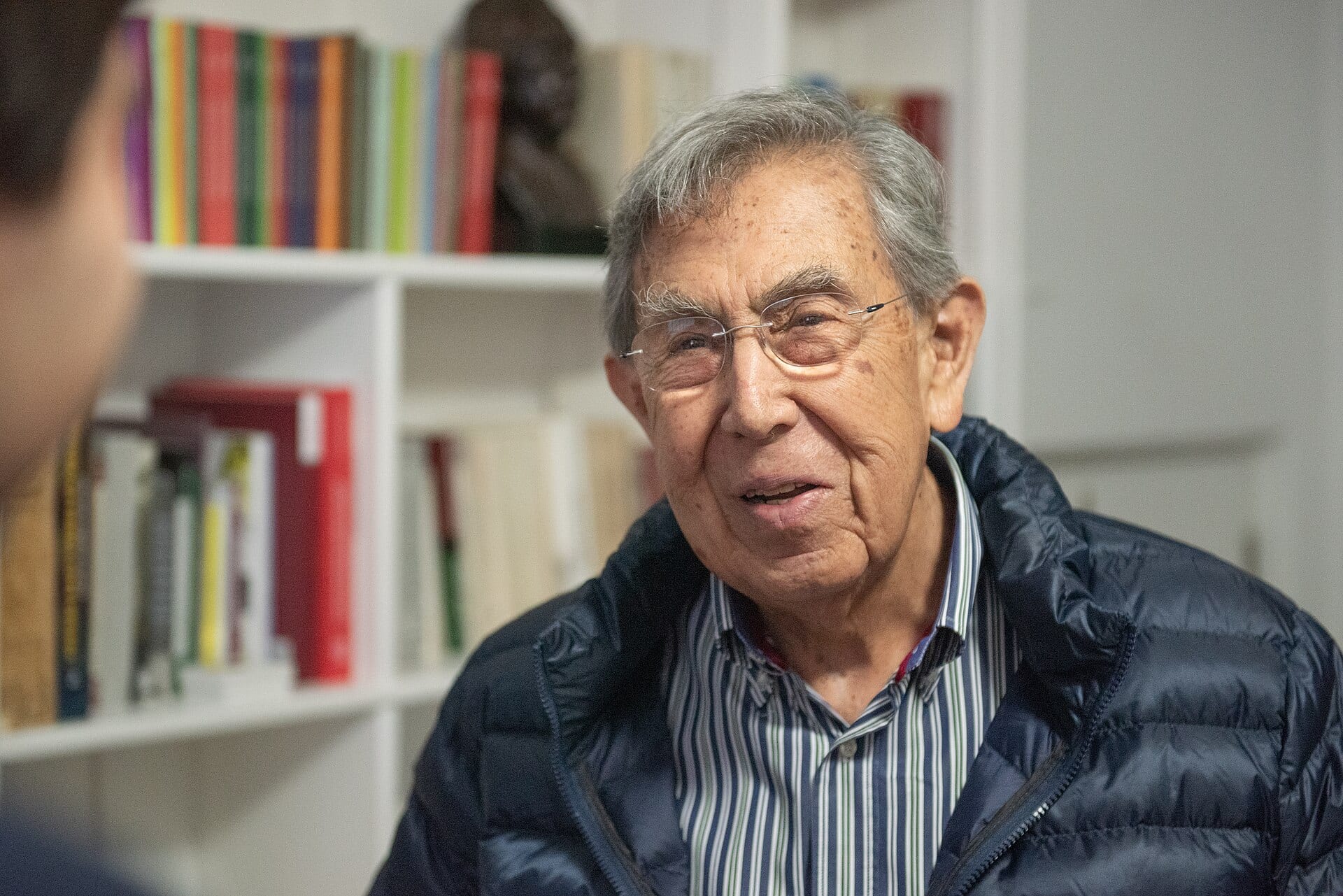 Cuauhtémoc Cárdenas, wearing glasses and a navy puffer jacket, speaks in front of bookshelves in a February 2022 portrait.