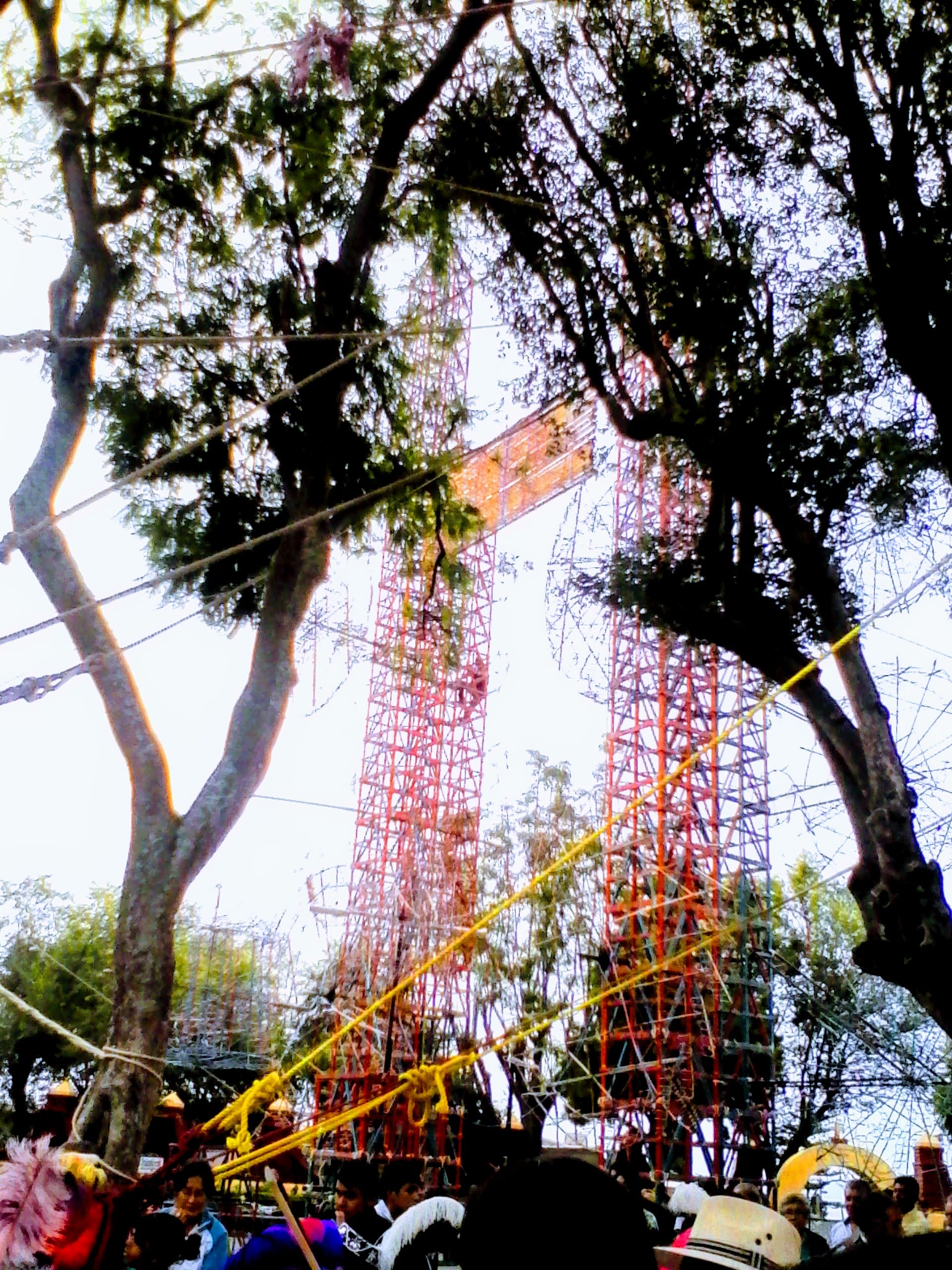 Dos castillos pirotécnicos de carrizo y cuerda se alzan entre árboles ante una multitud, en una fiesta patronal.