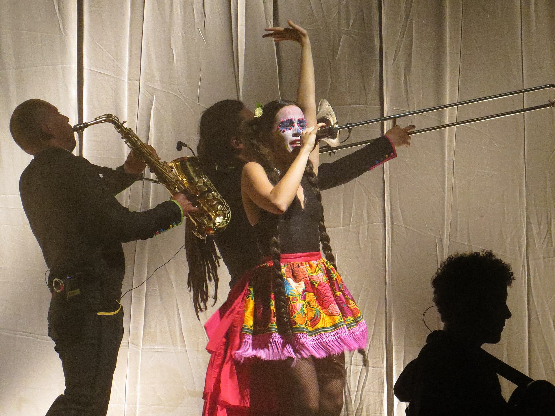 Una cantante con el rostro pintado de calavera levanta un brazo junto a dos músicos que tocan saxofón y trombón sobre un escenario.