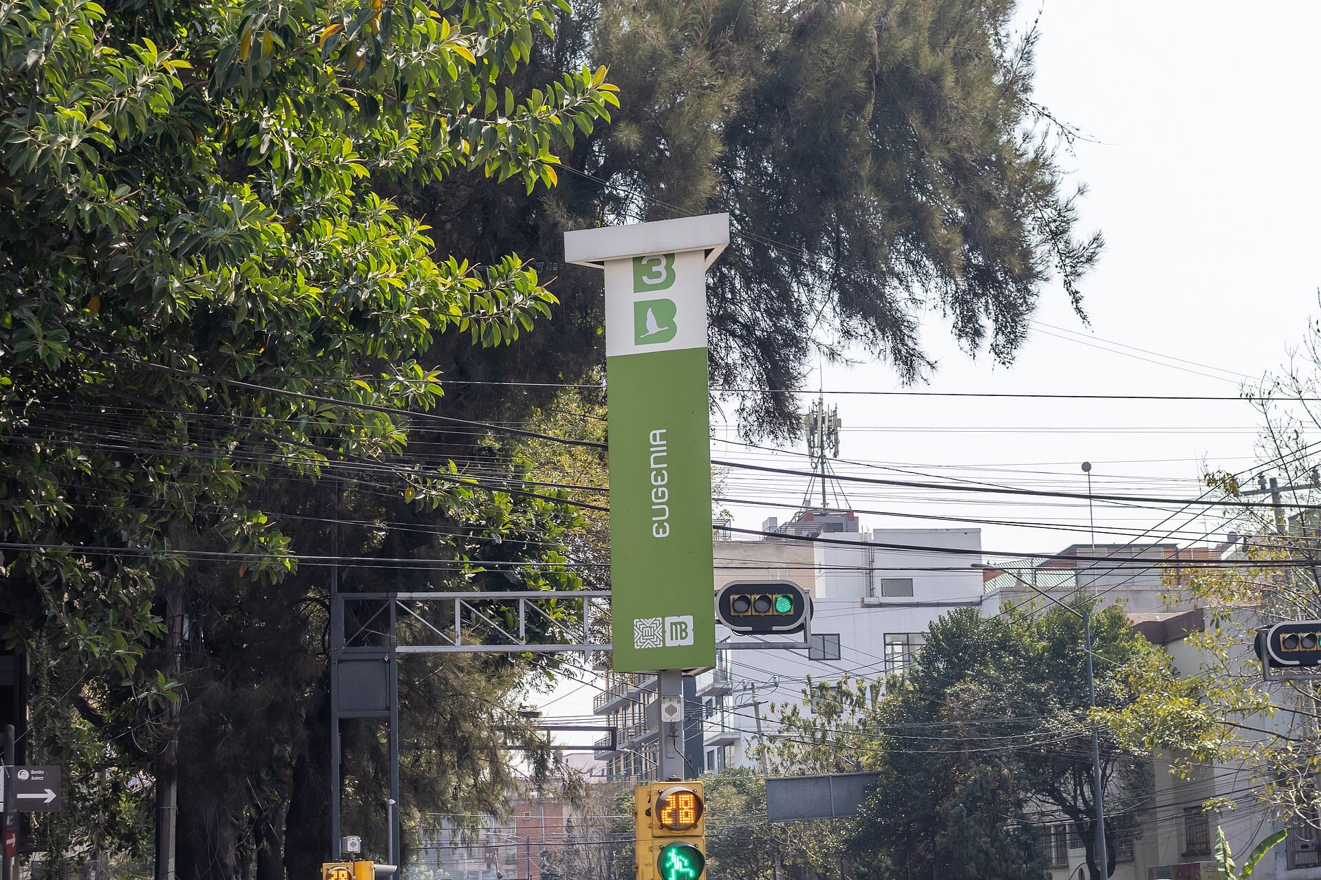 Poste verde y blanco de la estación Eugenia de la Línea 3 del Metrobús, junto a un semáforo, árboles y edificios bajos.