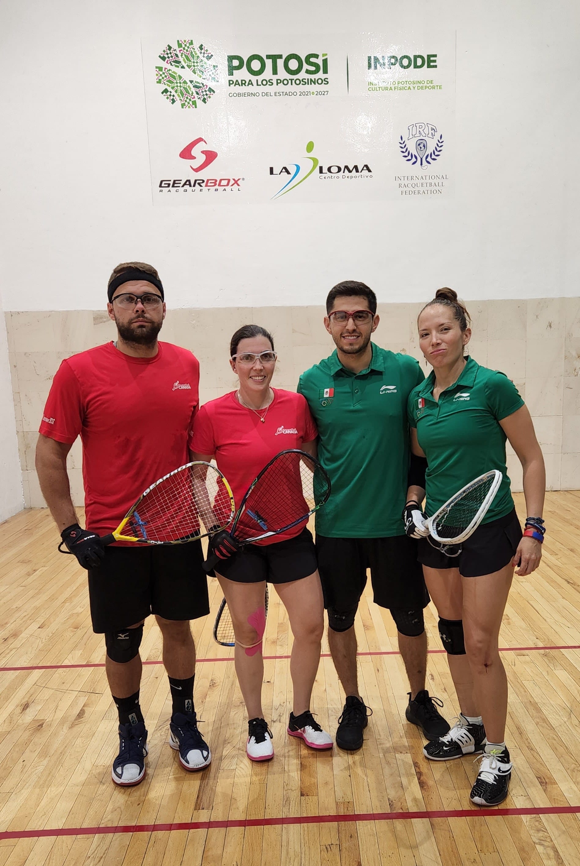 Dos jugadores canadienses con playera roja y dos mexicanos con polo verde posan con sus raquetas en una cancha de madera.