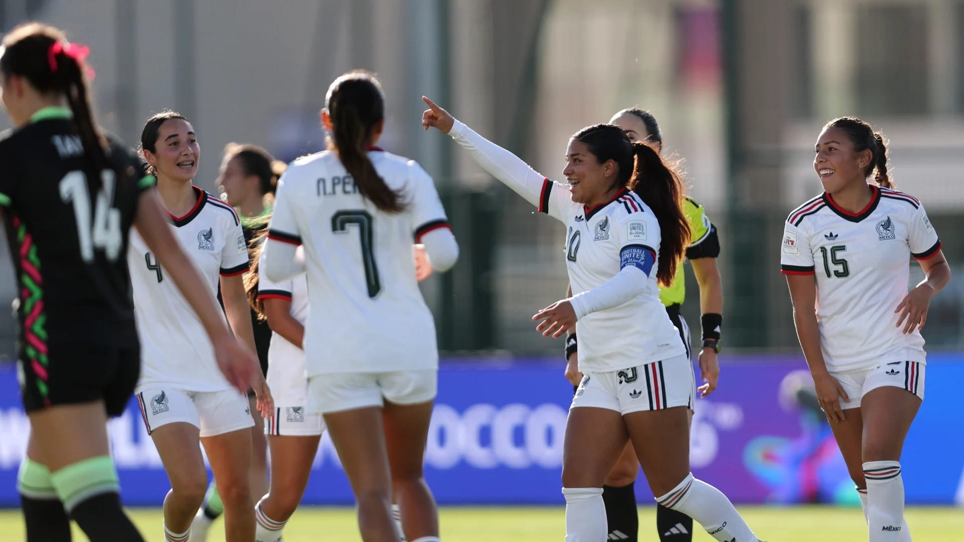 A Mexico player in a white jersey points ahead while teammates smile beside her after a goal.