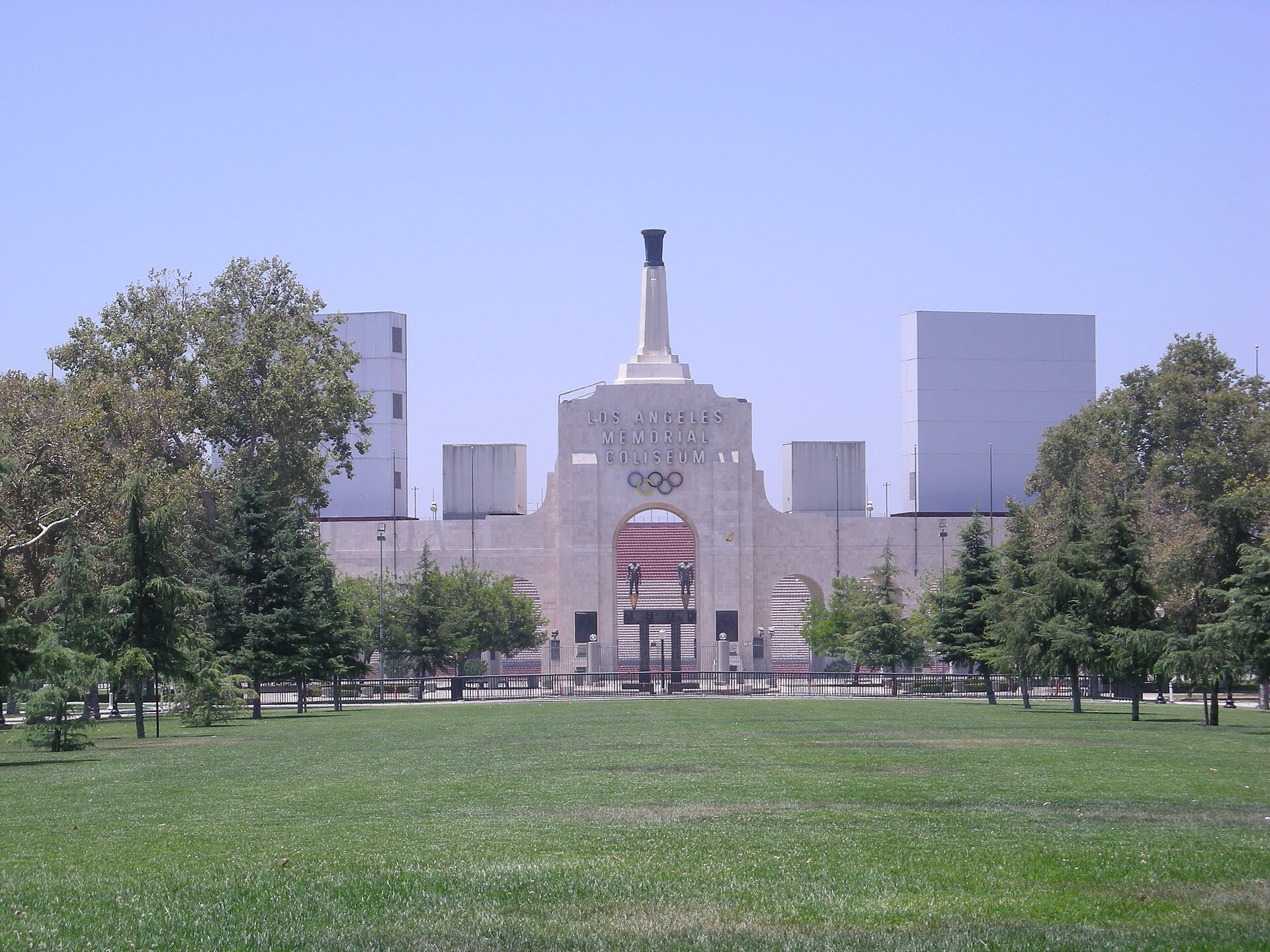 The pale stone facade of the Los Angeles Memorial Coliseum and its torch tower, seen from a grassy park with trees.