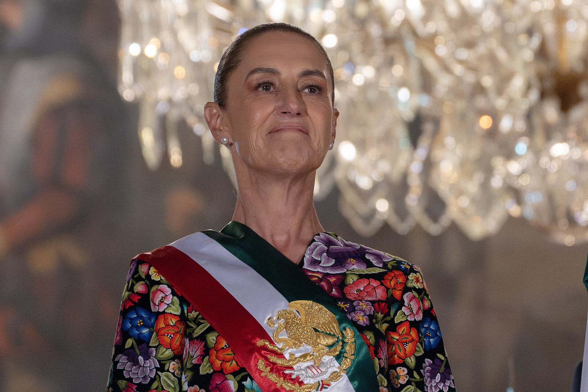 La presidenta Claudia Sheinbaum, con la banda presidencial sobre un traje bordado de flores, bajo una lámpara de cristal.