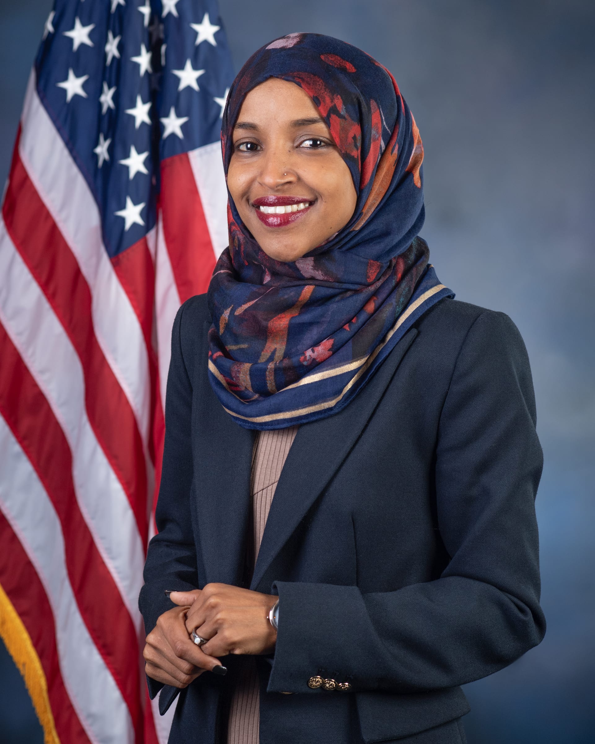 Retrato oficial de Ilhan Omar, con hiyab de color claro, saco oscuro y una bandera de Estados Unidos al fondo.