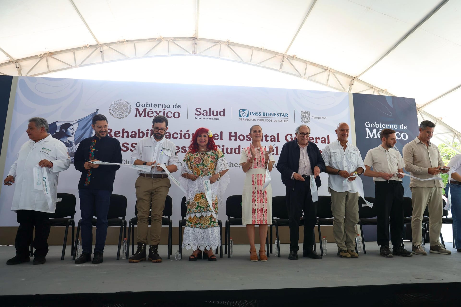 La presidenta Claudia Sheinbaum aplaude de pie junto a otras ocho personas que sostienen listones en un escenario con el letrero del Gobierno de México y de IMSS Bienestar.