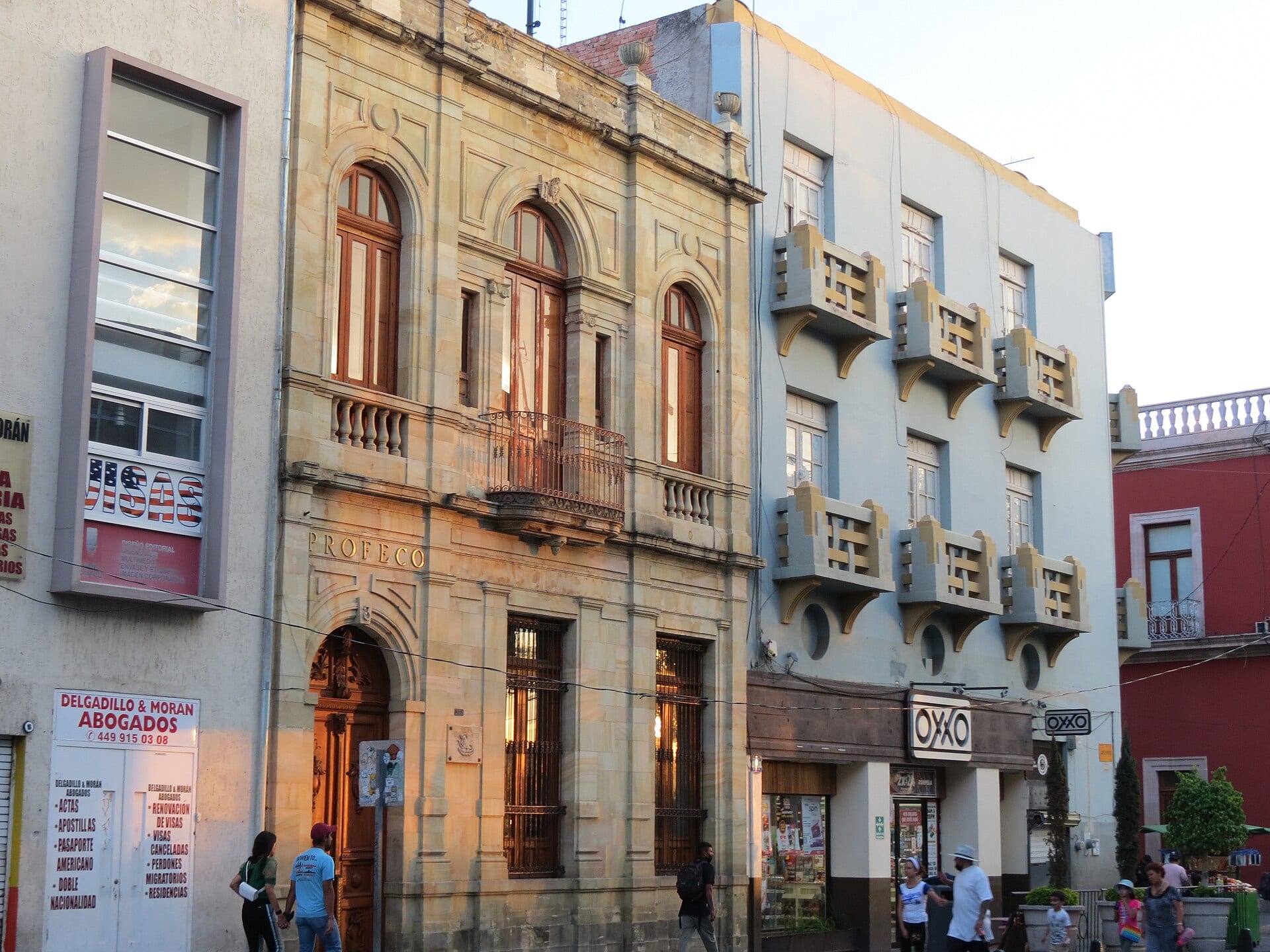 Fachada de piedra de dos pisos con la palabra Profeco sobre la puerta de arco, junto a un edificio con una tienda de conveniencia, con peatones en la banqueta.
