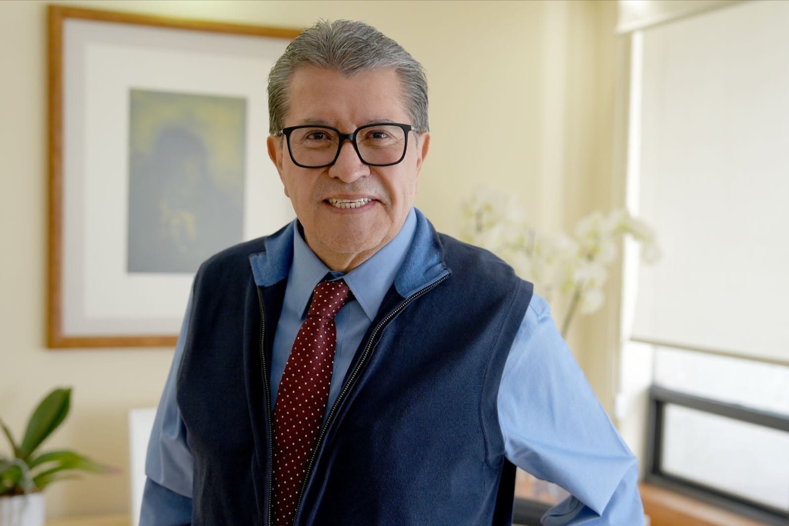 Ricardo Monreal, con lentes de armazón negro, chaleco azul marino y corbata roja de puntos, sonríe a la cámara en una oficina.