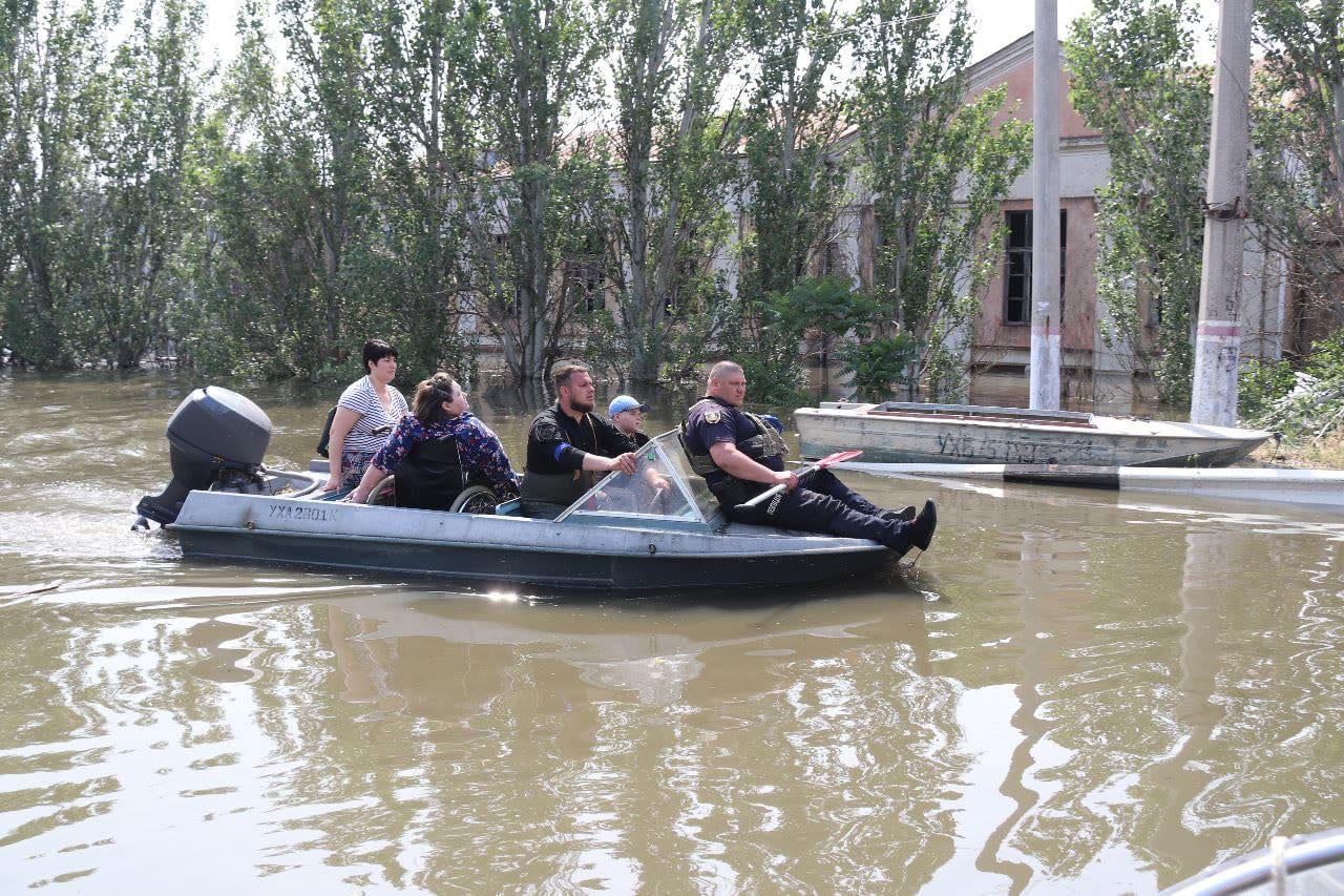 Una lancha con cinco personas, entre ellas un policía con remo y una mujer en silla de ruedas, avanza por una calle inundada entre árboles y edificios.