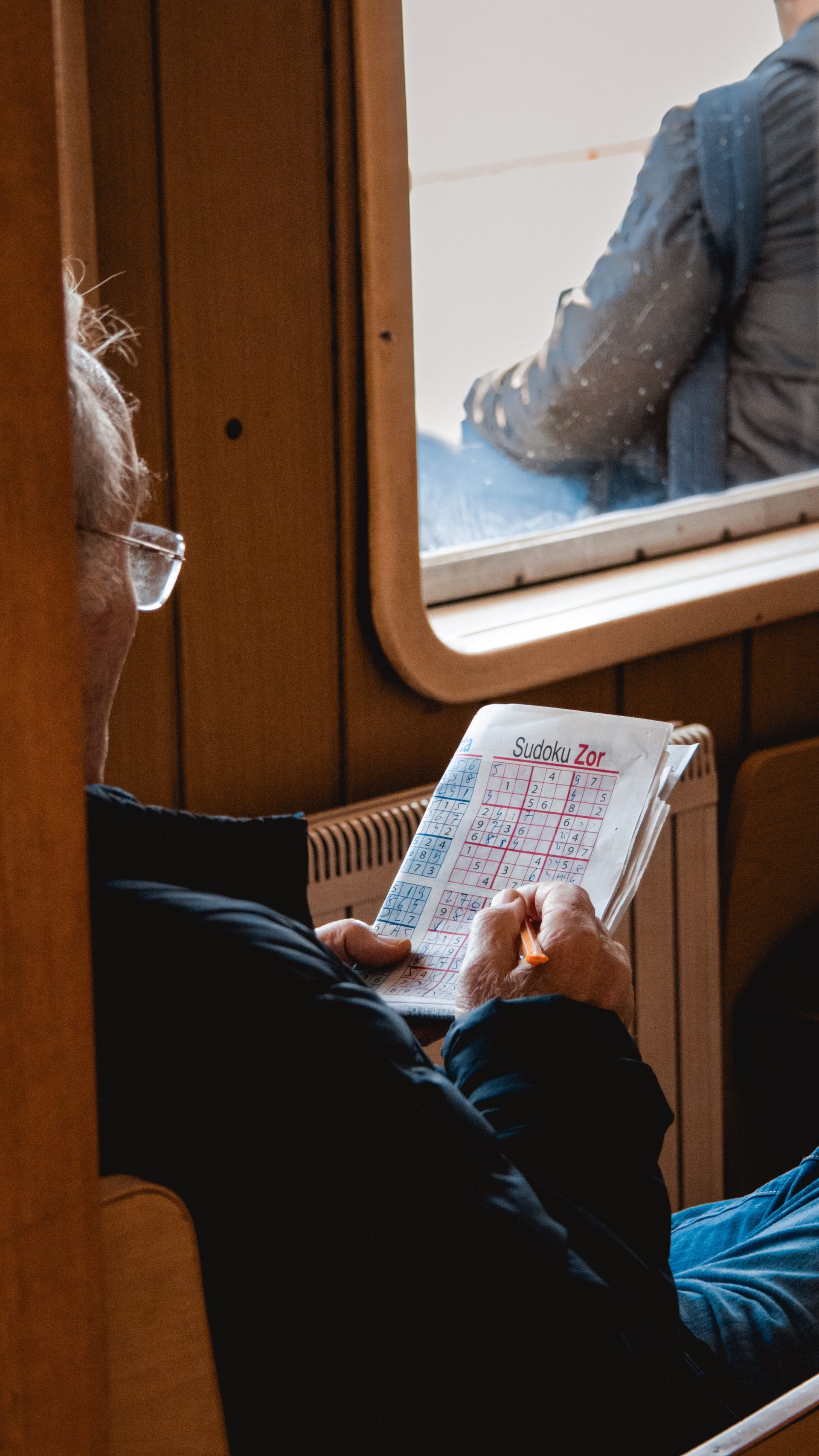 Un hombre mayor sentado junto a una ventana concentrado resolviendo un sudoku en papel.