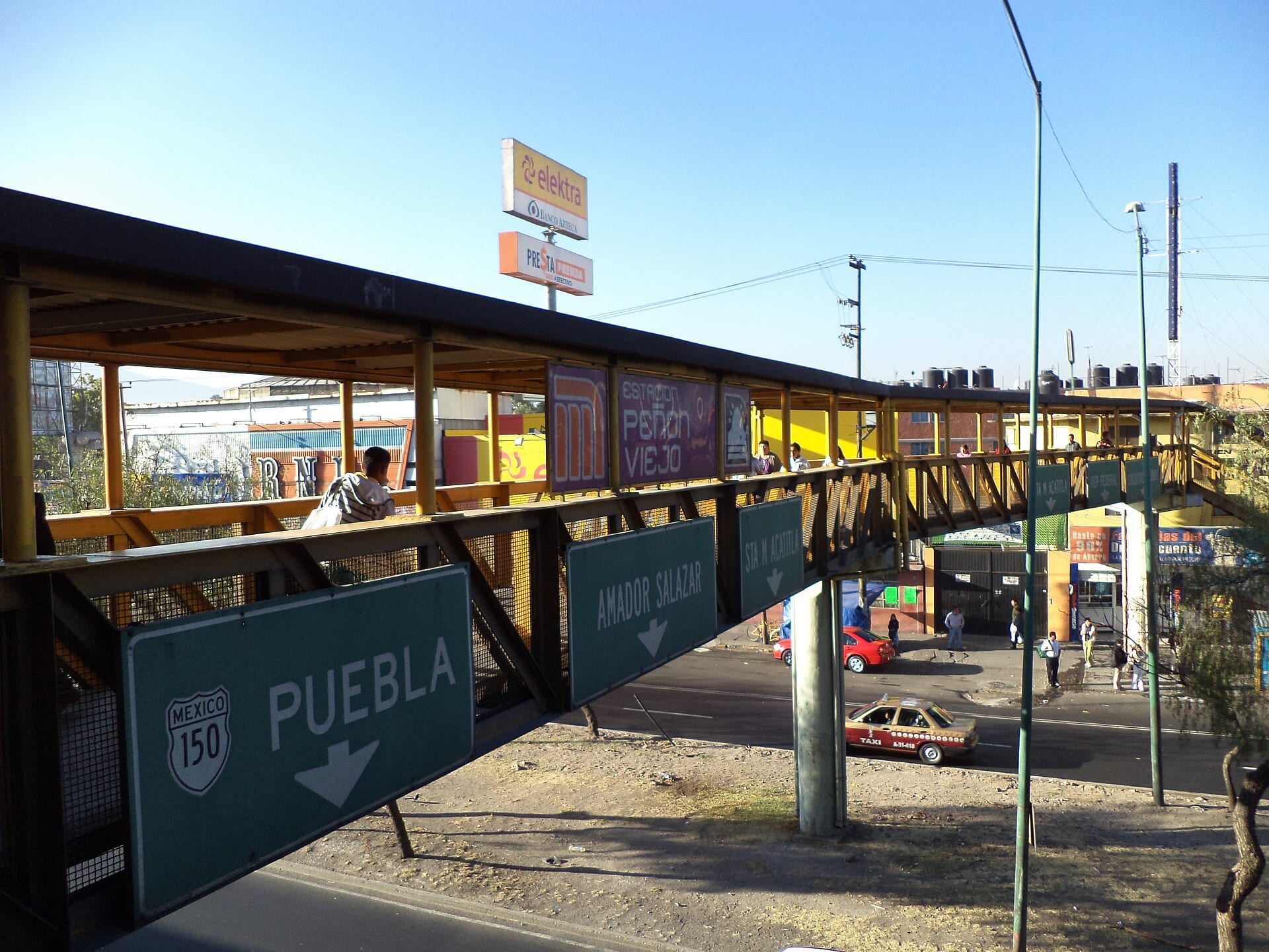 Puente peatonal amarillo con el letrero de la estación Peñón Viejo sobre una avenida con señales de tránsito.
