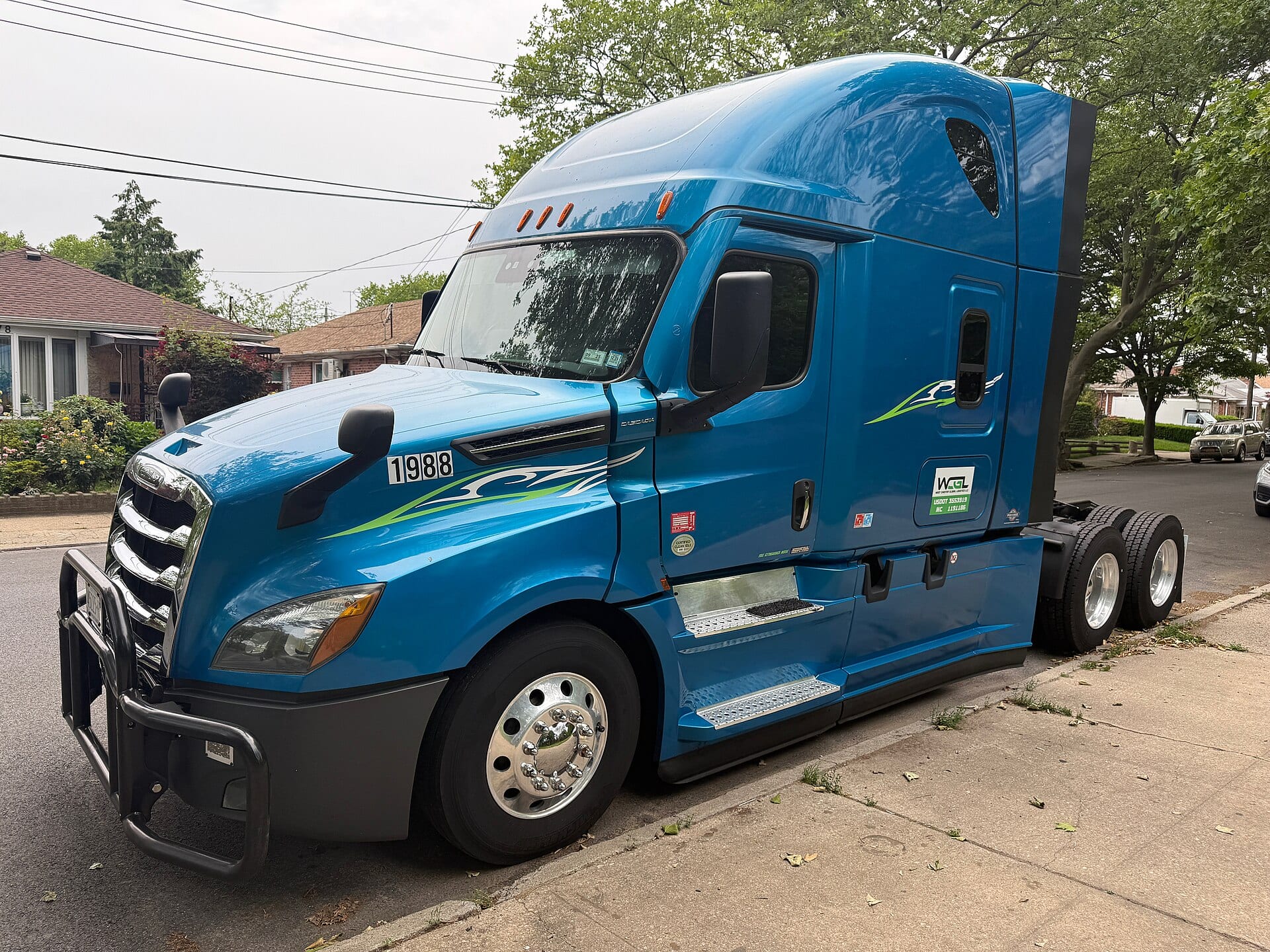 Un tractocamión Freightliner Cascadia azul estacionado junto a la banqueta de una calle residencial con árboles.