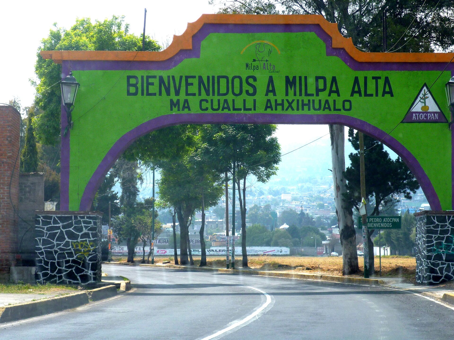Arco verde y morado con el letrero de bienvenida a Milpa Alta sobre una carretera, con árboles y casas al fondo.