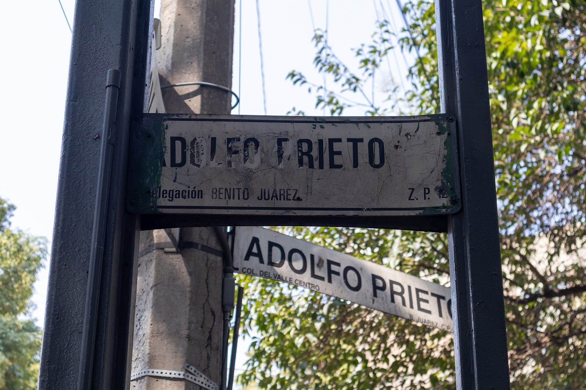 Placa de nomenclatura de la calle Adolfo Prieto, en la delegación Benito Juárez, junto a un poste y ramas de árbol.