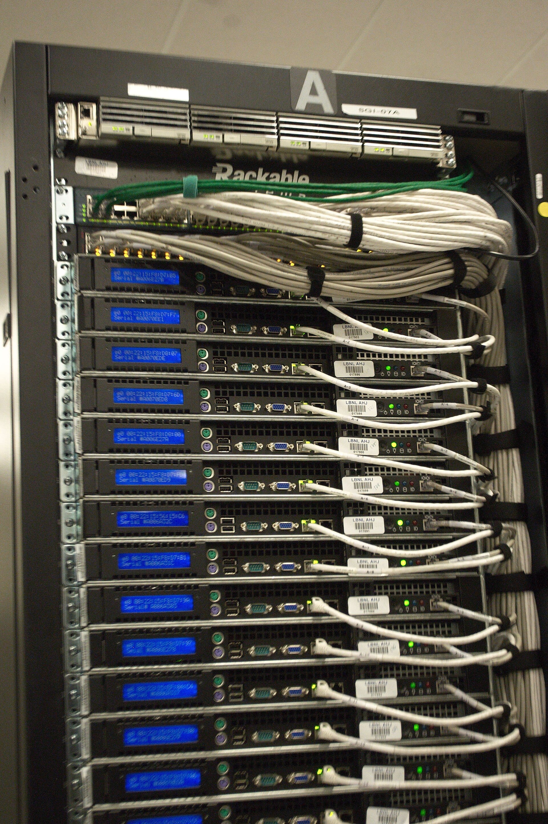 Close-up of a server cabinet with stacked equipment, small blue displays and white network cables bundled with ties.