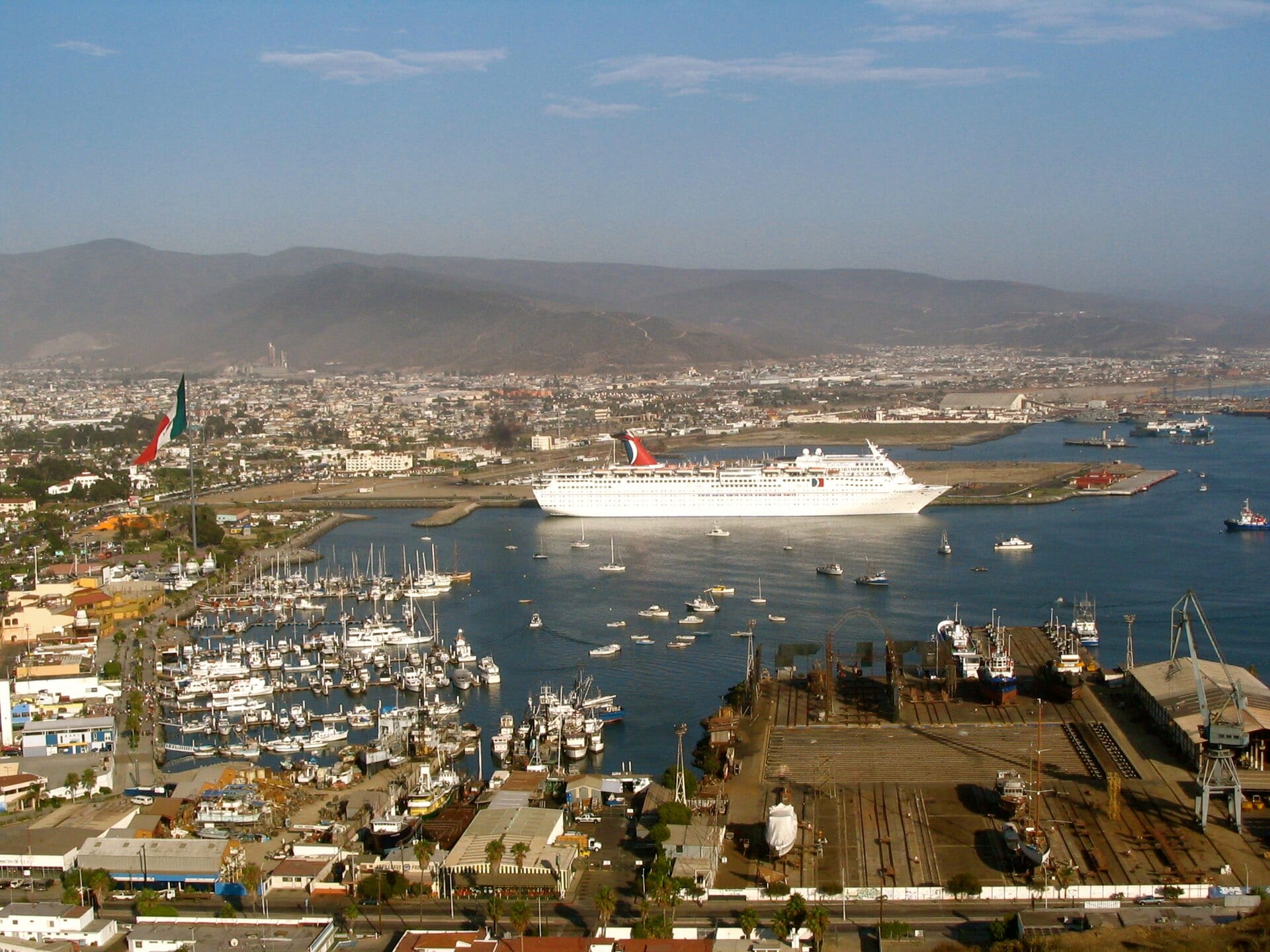 Aerial view of the port of Ensenada with a white cruise ship in the bay, sailboats in the marina and hills in the background.
