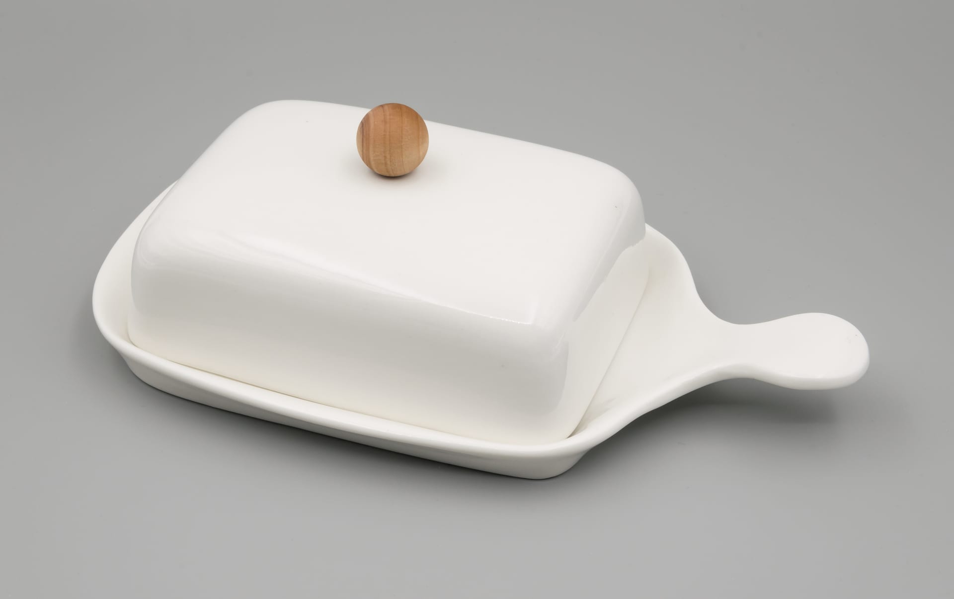 Una mantequillera de cerámica blanca con tapa y una perilla de madera, sobre un fondo gris liso.
