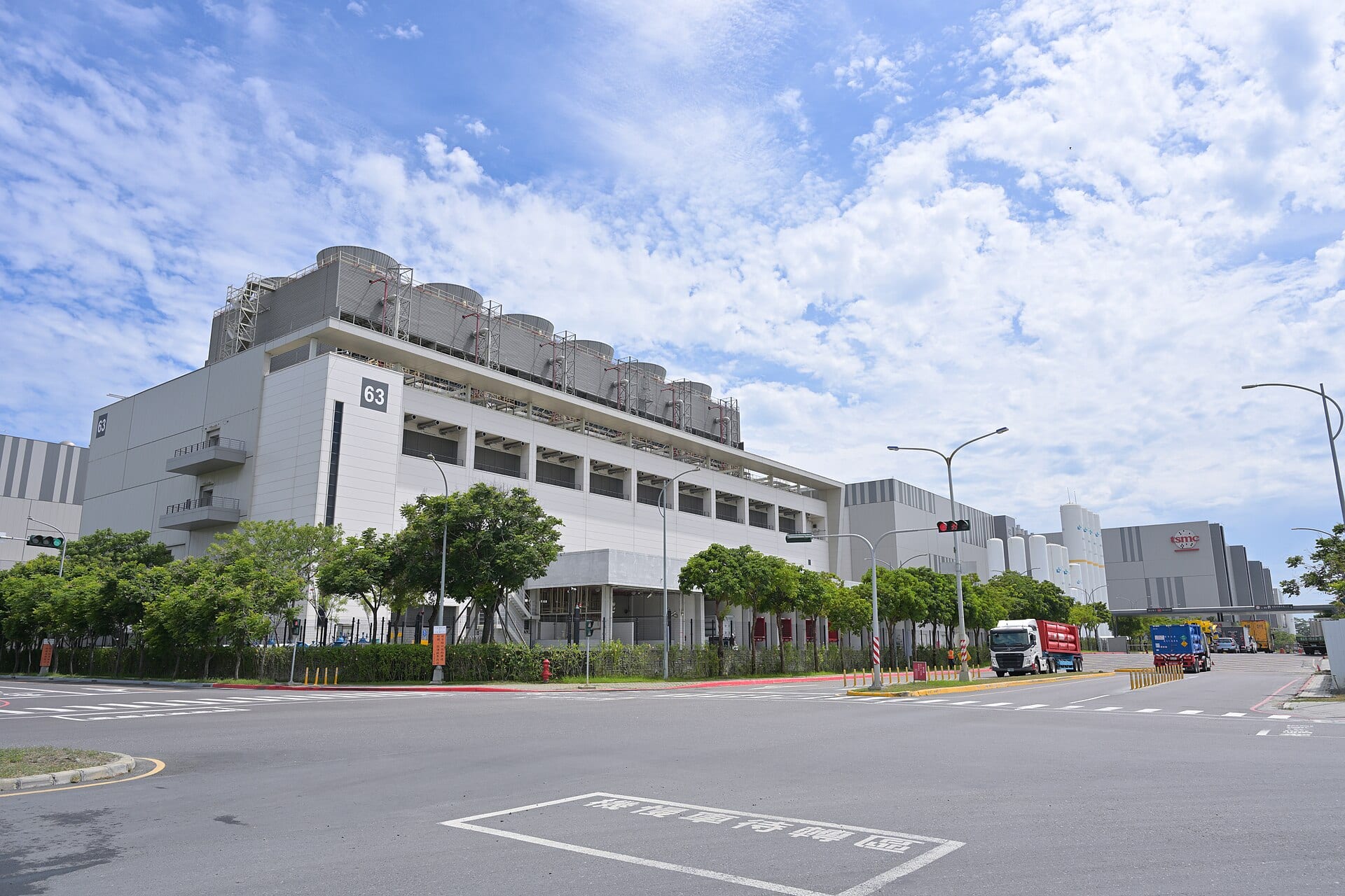 Edificio industrial blanco y gris de una planta de TSMC con torres de enfriamiento en el techo, junto a una calle con camiones.