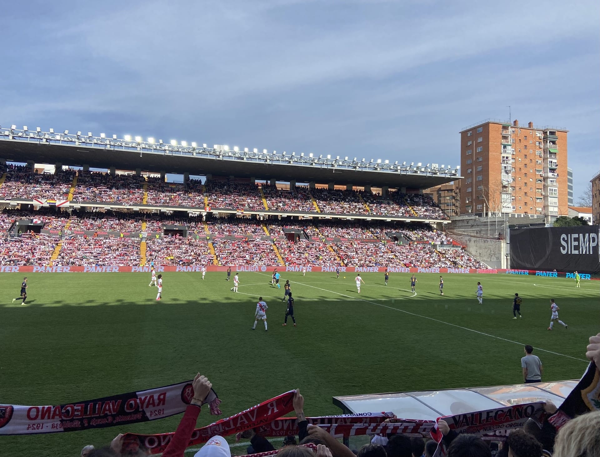 Gradas llenas del Estadio de Vallecas durante un partido, con aficionados que alzan bufandas del Rayo Vallecano.