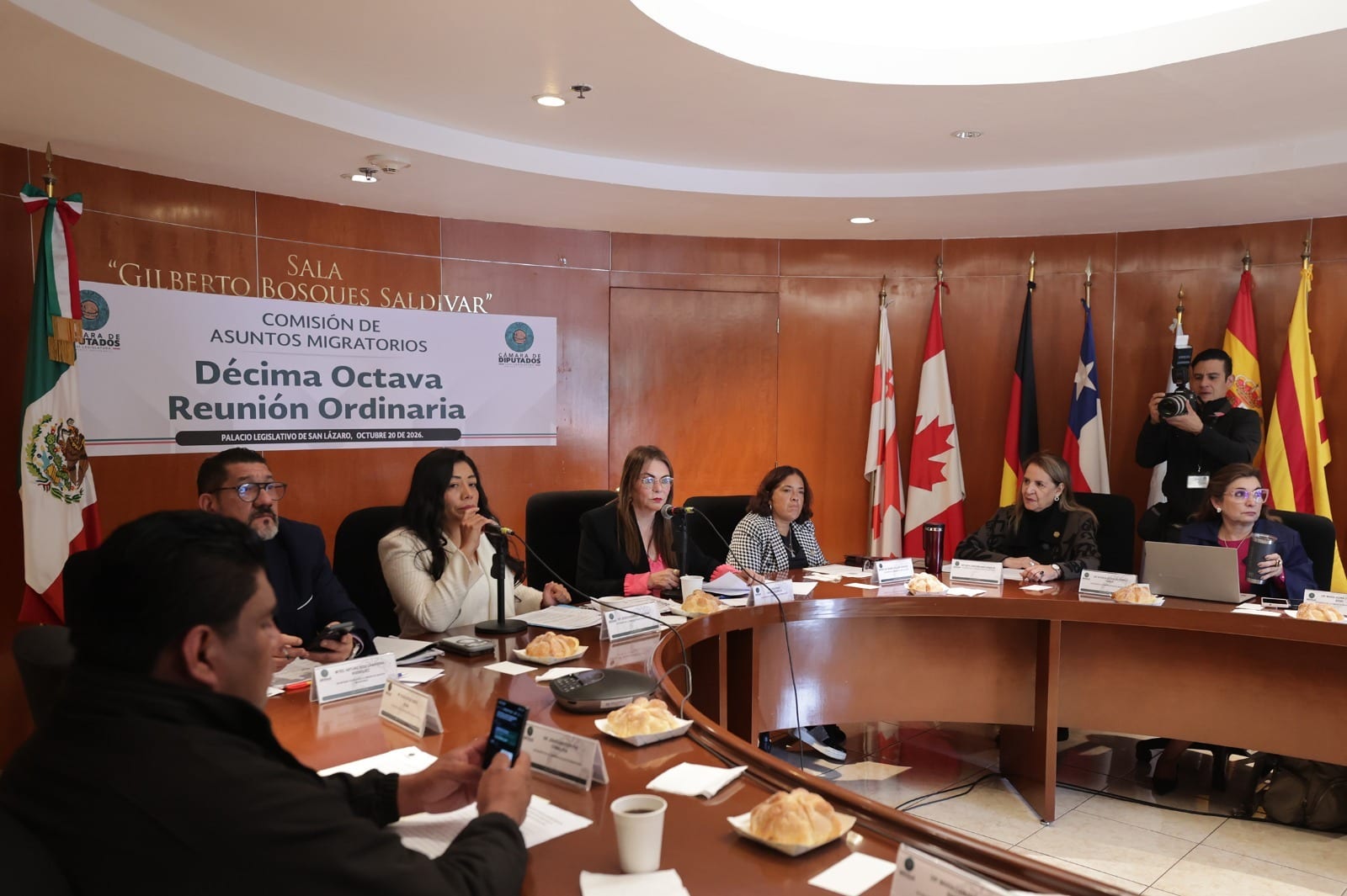 Legisladoras y legisladores sentados a una mesa curva bajo un cartel de la Décima Octava Reunión Ordinaria de la Comisión de Asuntos Migratorios, con banderas al fondo.