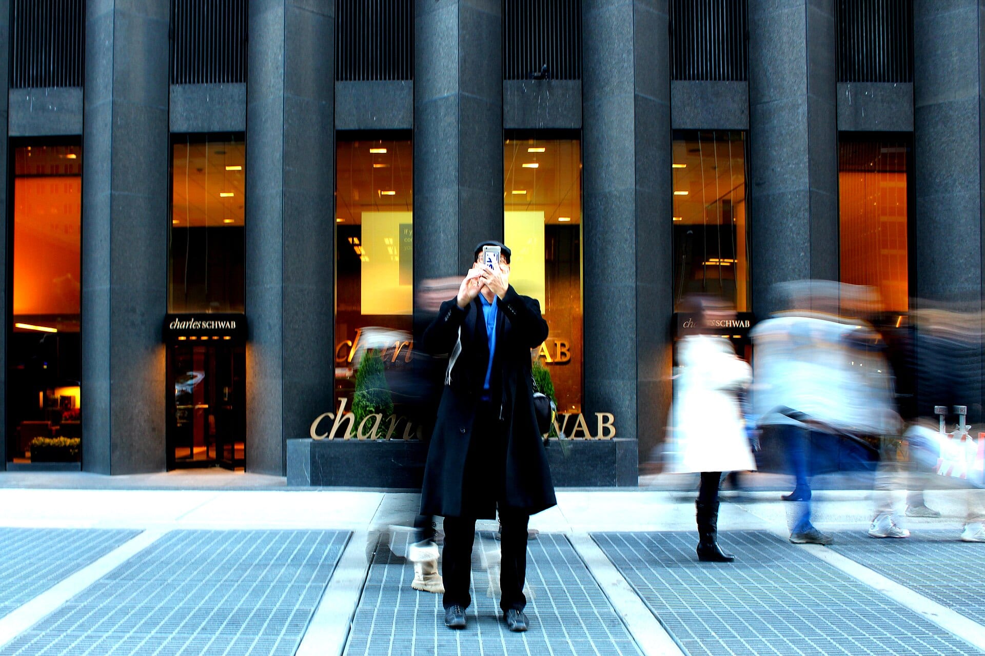 A man in a dark coat photographs with his phone in front of a Charles Schwab branch, while passersby blur past.