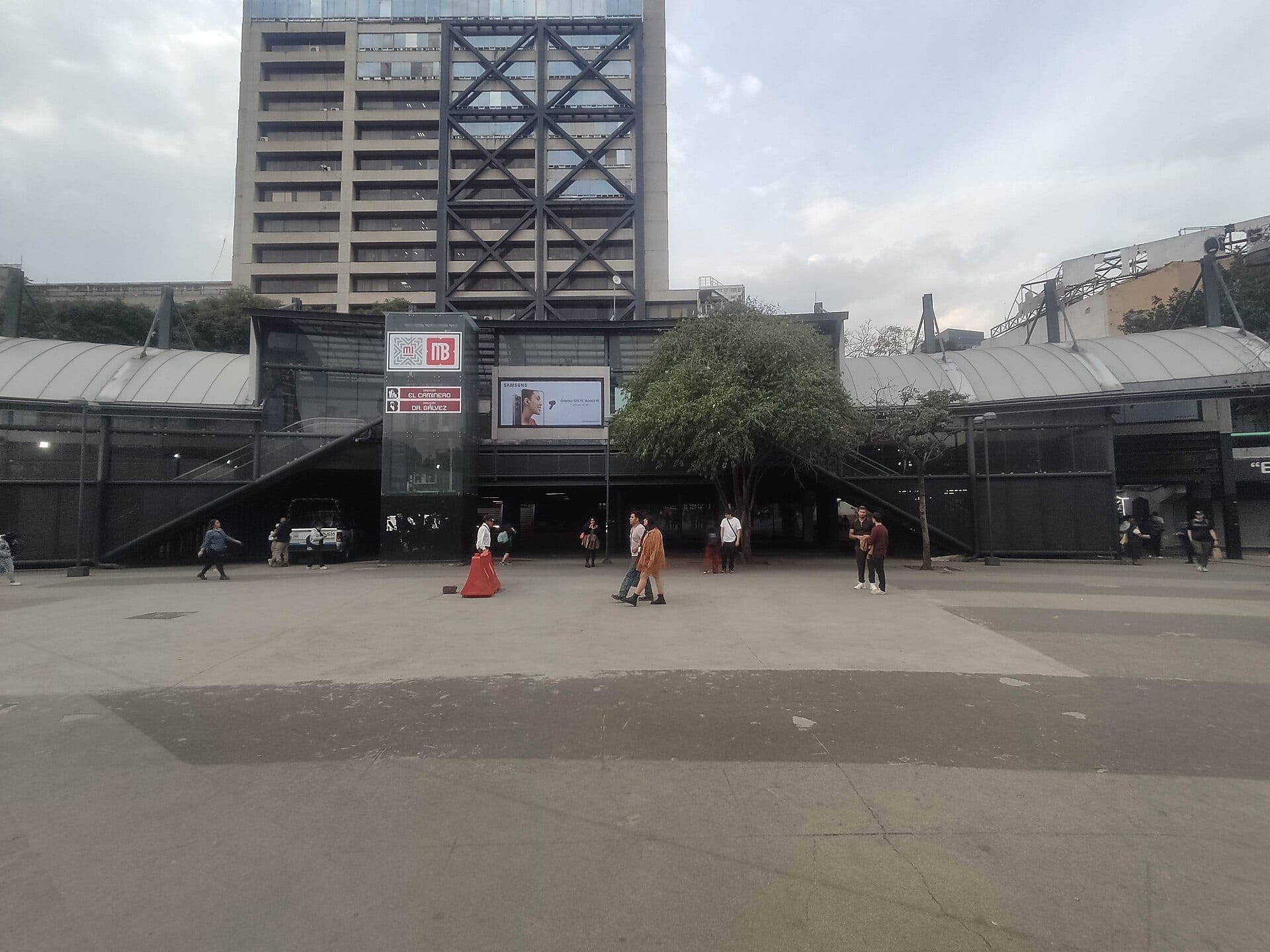 Fachada de una estación del Metrobús con los letreros de dirección El Caminero y Doctor Gálvez, y peatones cruzando la explanada frente a ella.