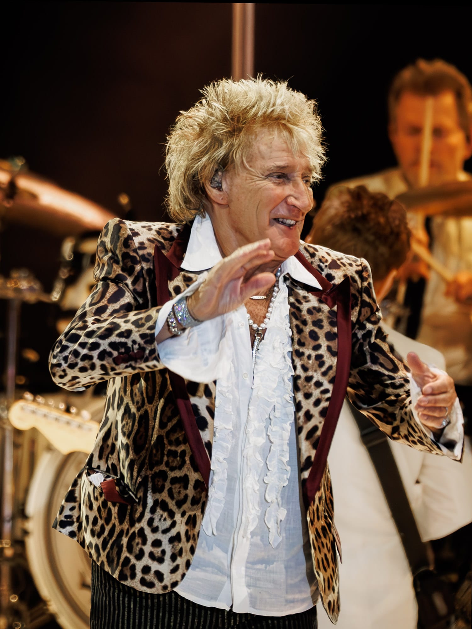 Rod Stewart, con saco de estampado de leopardo y camisa blanca de holanes, levanta una mano sobre el escenario durante un concierto.