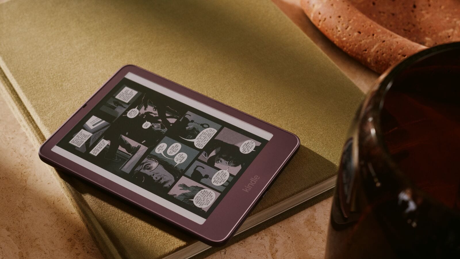 Un lector Kindle de borde morado sobre un libro de tapa verde muestra una página de cómic a color; junto a él hay una copa oscura y una pieza de cerámica.