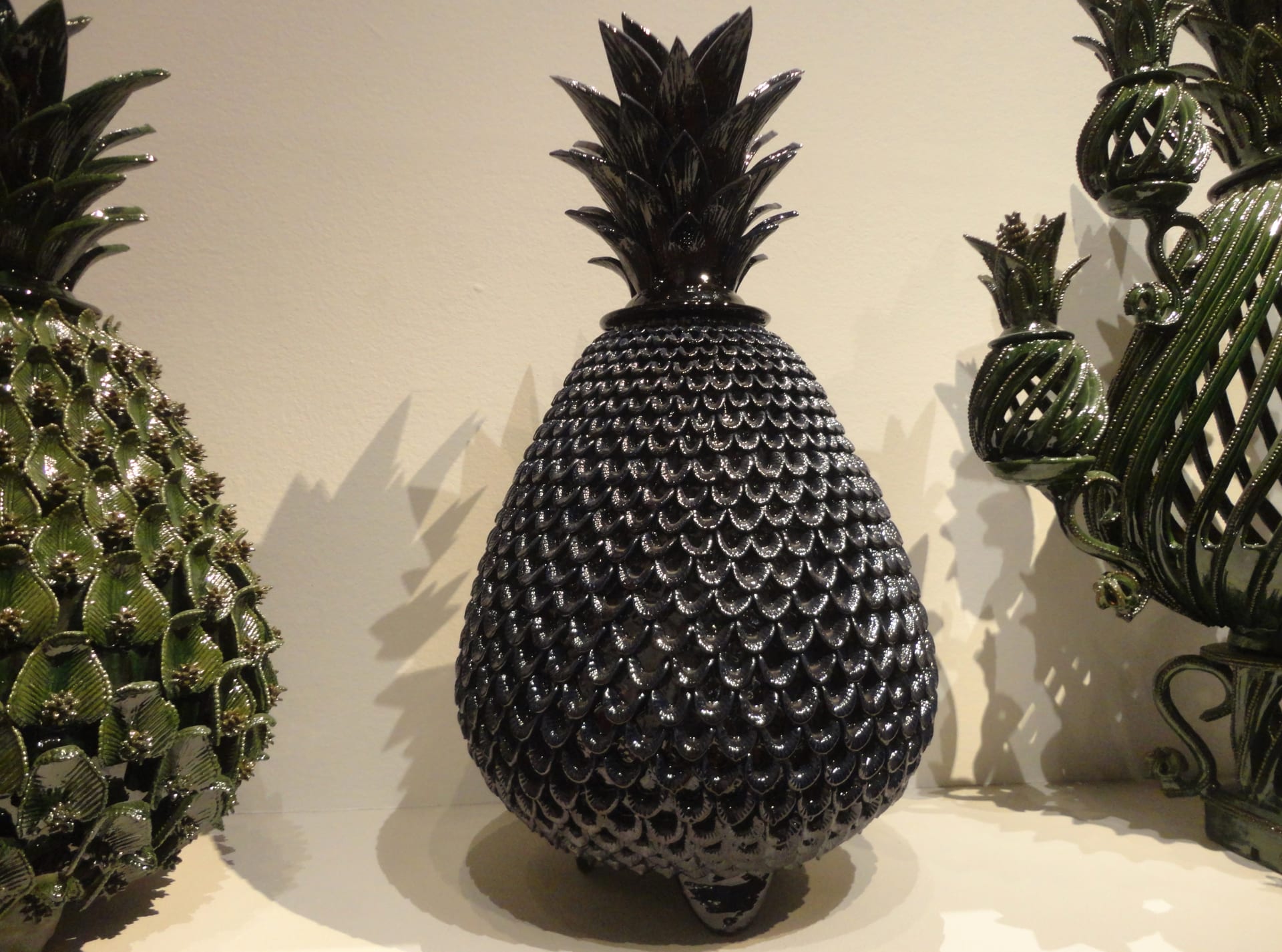 Piña decorativa de cerámica vidriada de tono negro azulado, con hojas puntiagudas, junto a otras piezas de barro verdes.