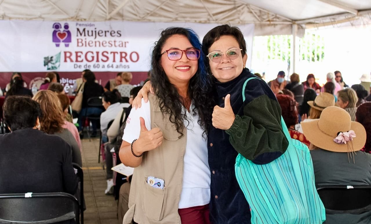 Dos mujeres sonríen con el pulgar arriba ante un módulo de registro con una manta de Pensión Mujeres Bienestar, de 60 a 64 años.