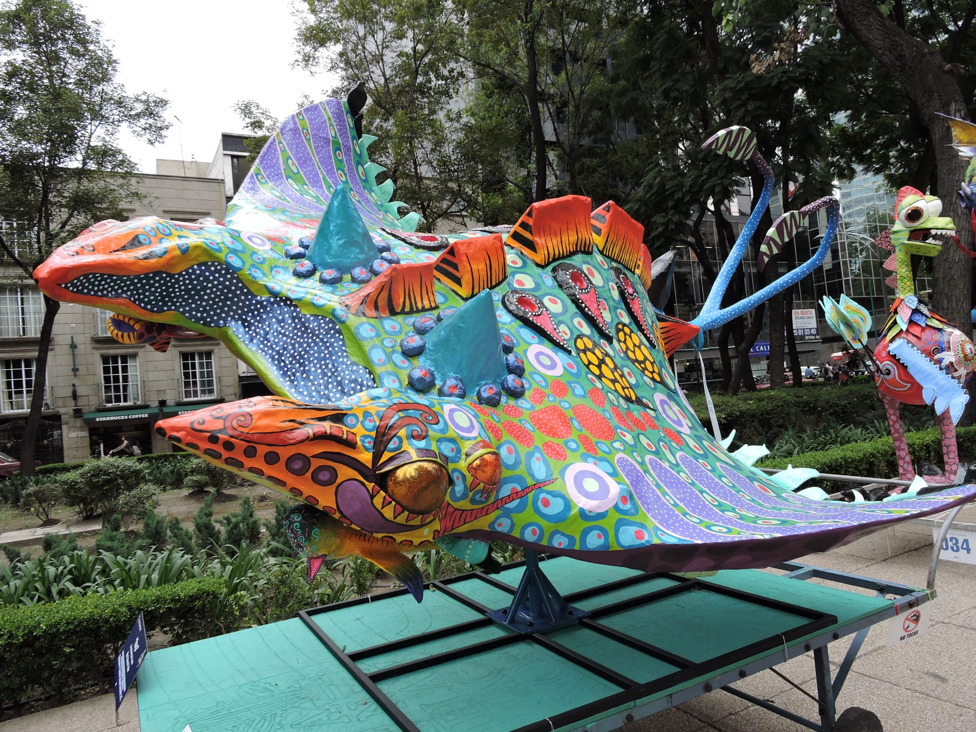 A giant stingray-shaped alebrije painted green, purple and orange sits on a wheeled platform beside trees and planters on an avenue.