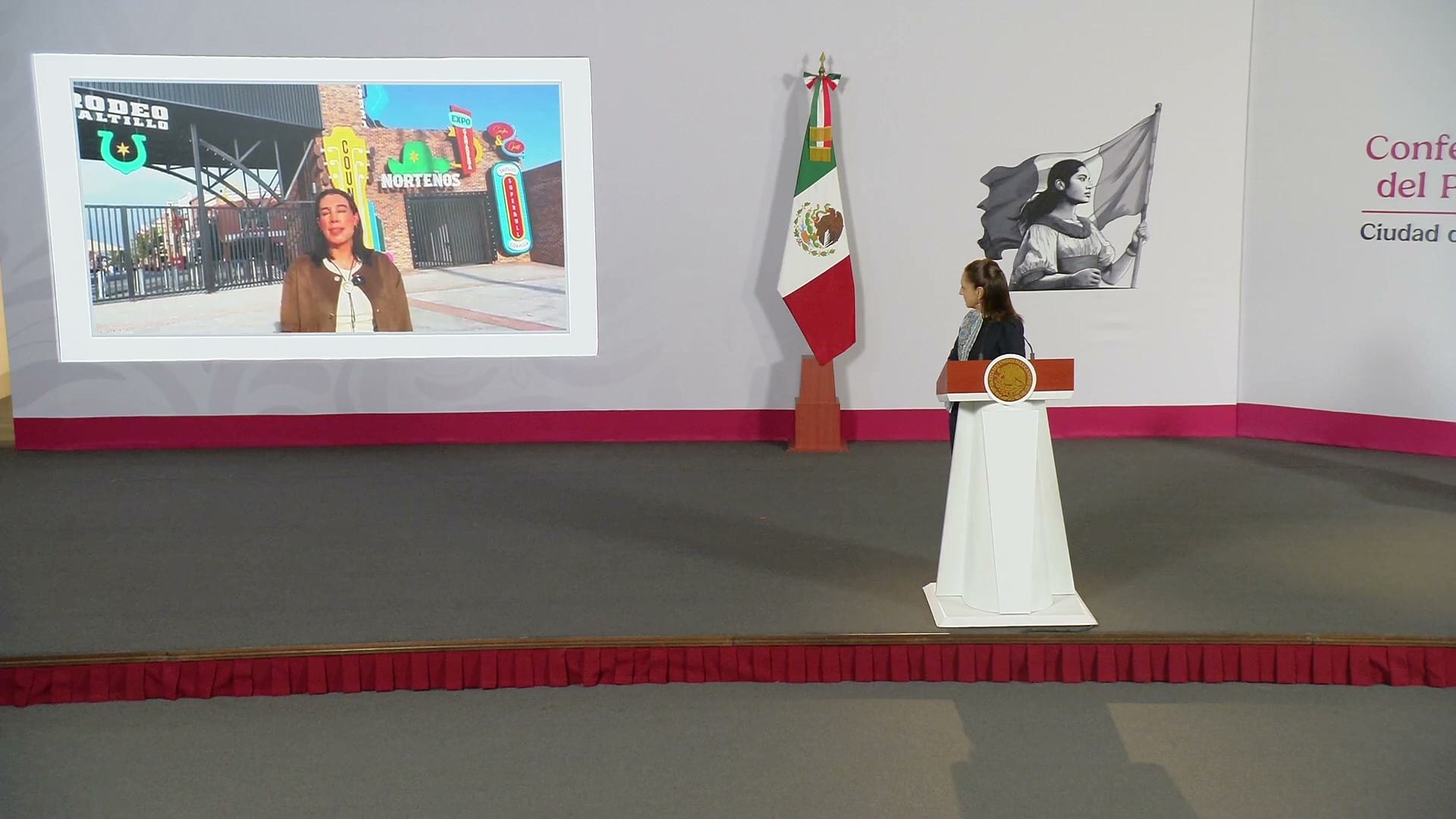 Una mujer aparece en una pantalla frente a un rodeo, junto al atril del salón de la conferencia matutina.