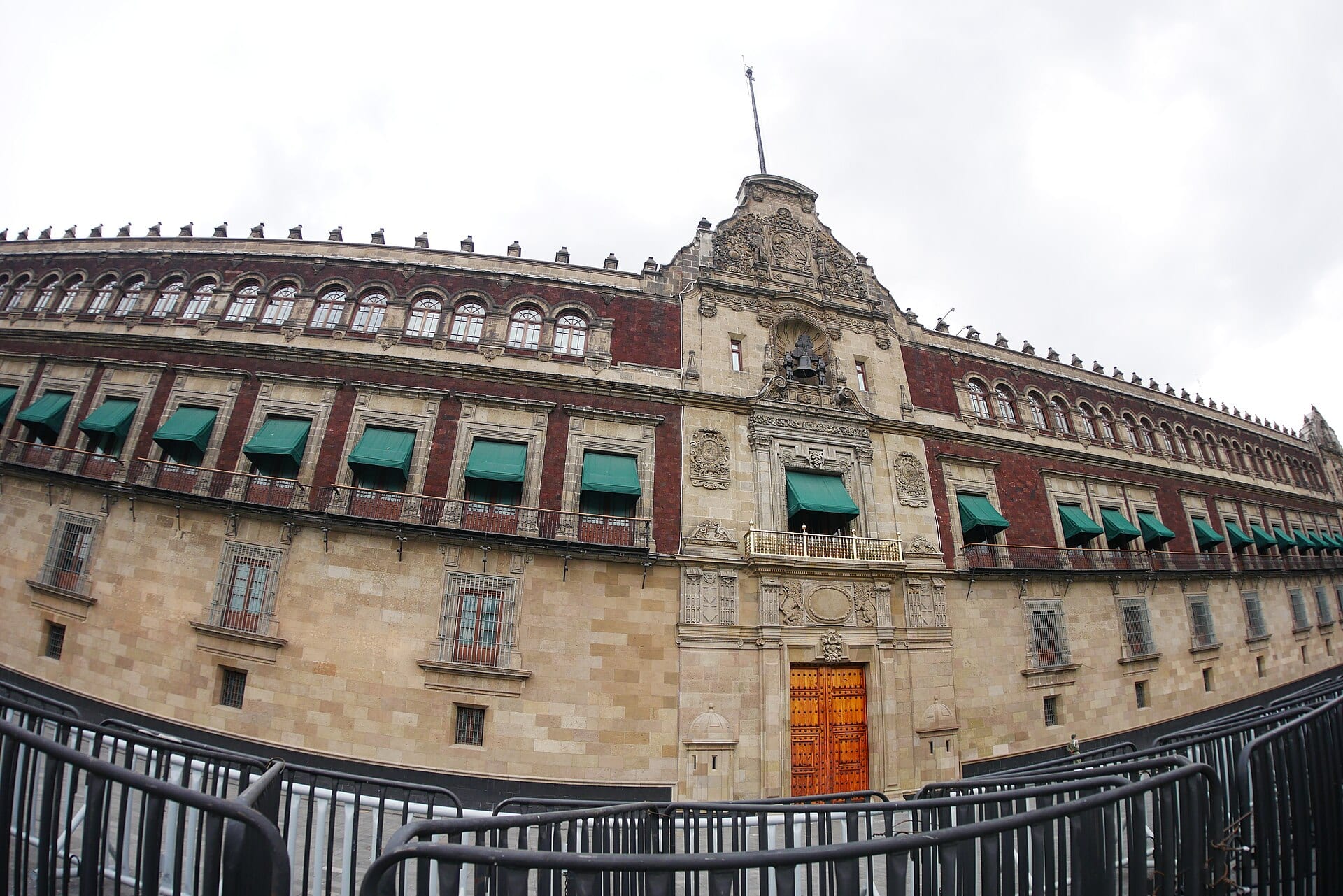 Fachada de Palacio Nacional con su portón de madera, el balcón central dorado y toldos verdes, detrás de unas vallas metálicas.